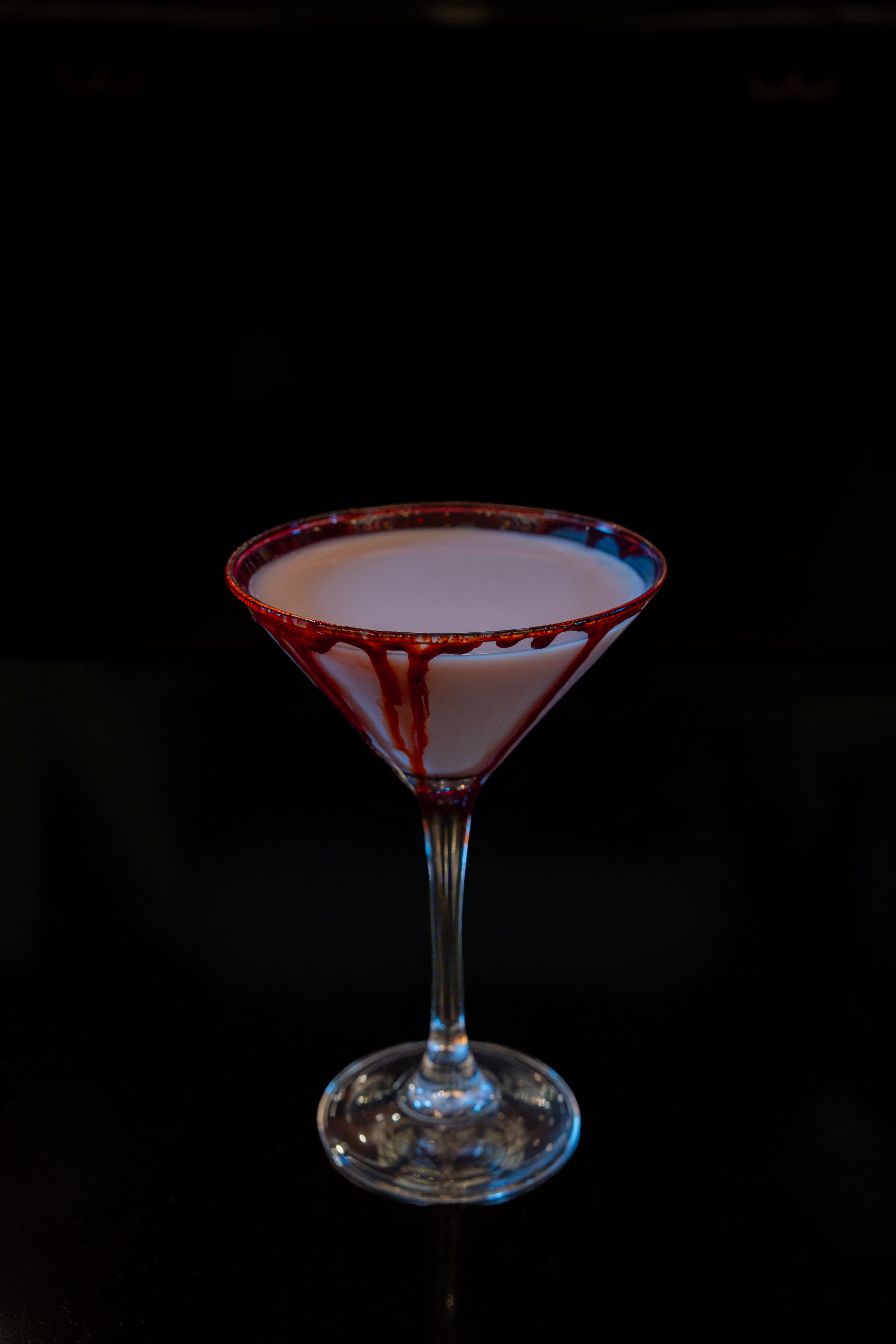 Un verre à martini contenant une boisson claire, dont le bord est nappé de sirop rouge qui coule le long de la paroi, sur fond sombre.