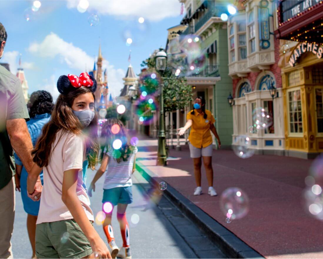 A girl wearing Minnie Mouse ears and a face mask walks down a lively, bubble-filled street at a theme park, accompanied by family and other visitors under bright, sunny weather.