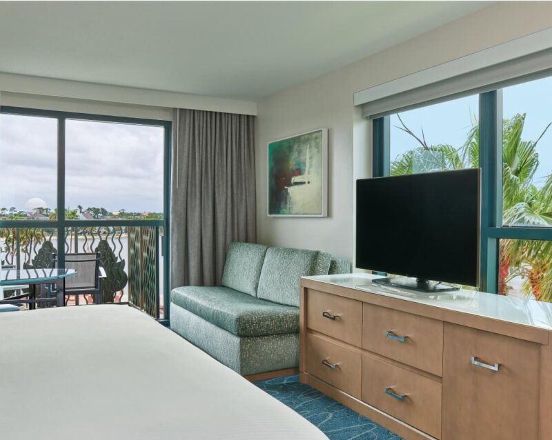 A hotel room with a bed, green sofa, flat-screen TV on a wooden dresser, large window with palm tree view, and balcony with chairs. Light walls, blue carpet, and framed art on the wall.