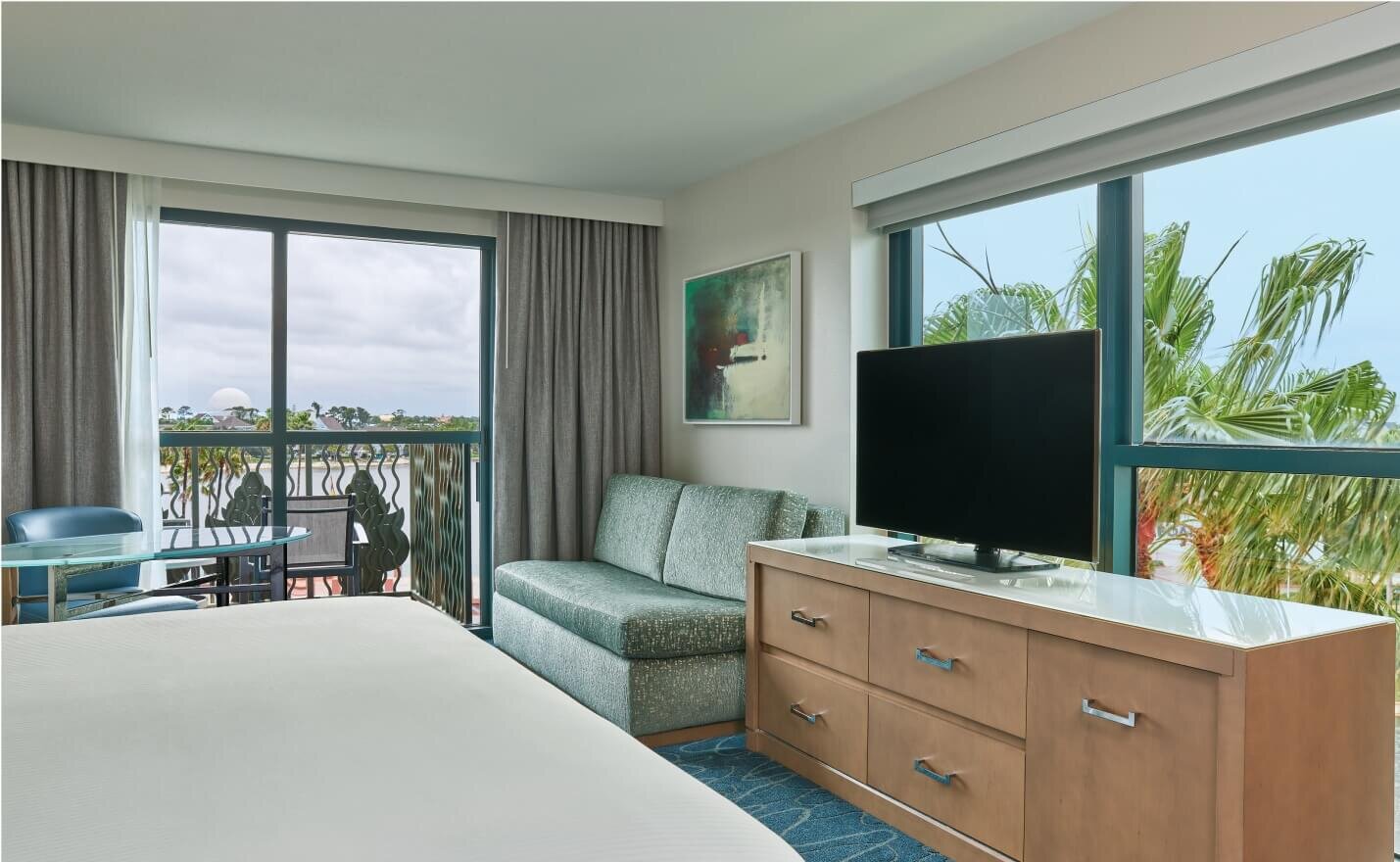 A hotel room with a bed, green sofa, flat-screen TV on a wooden dresser, large window with palm tree view, and balcony with chairs. Light walls, blue carpet, and framed art on the wall.