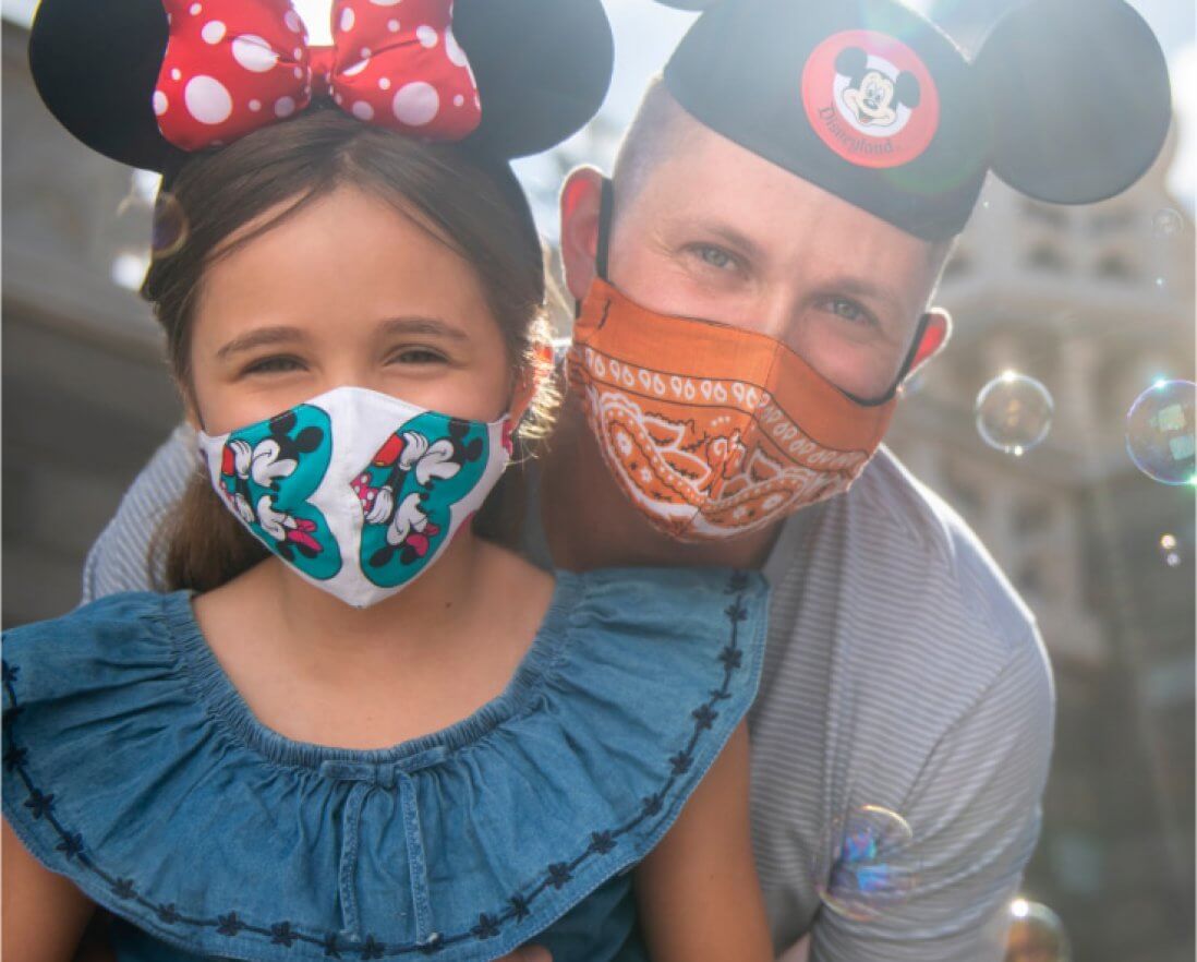 A man and young girl wear Mickey Mouse ear hats and face masks, with the girl in a Minnie Mouse bow and a blue dress. They smile at the camera outdoors, with bubbles floating around them.