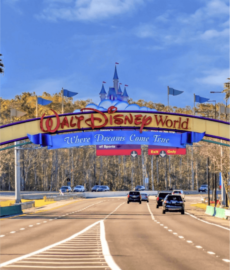 Cars drive under the colorful Walt Disney World entrance arch, which features a blue castle and the slogan Where Dreams Come True, against a backdrop of trees and a clear sky.