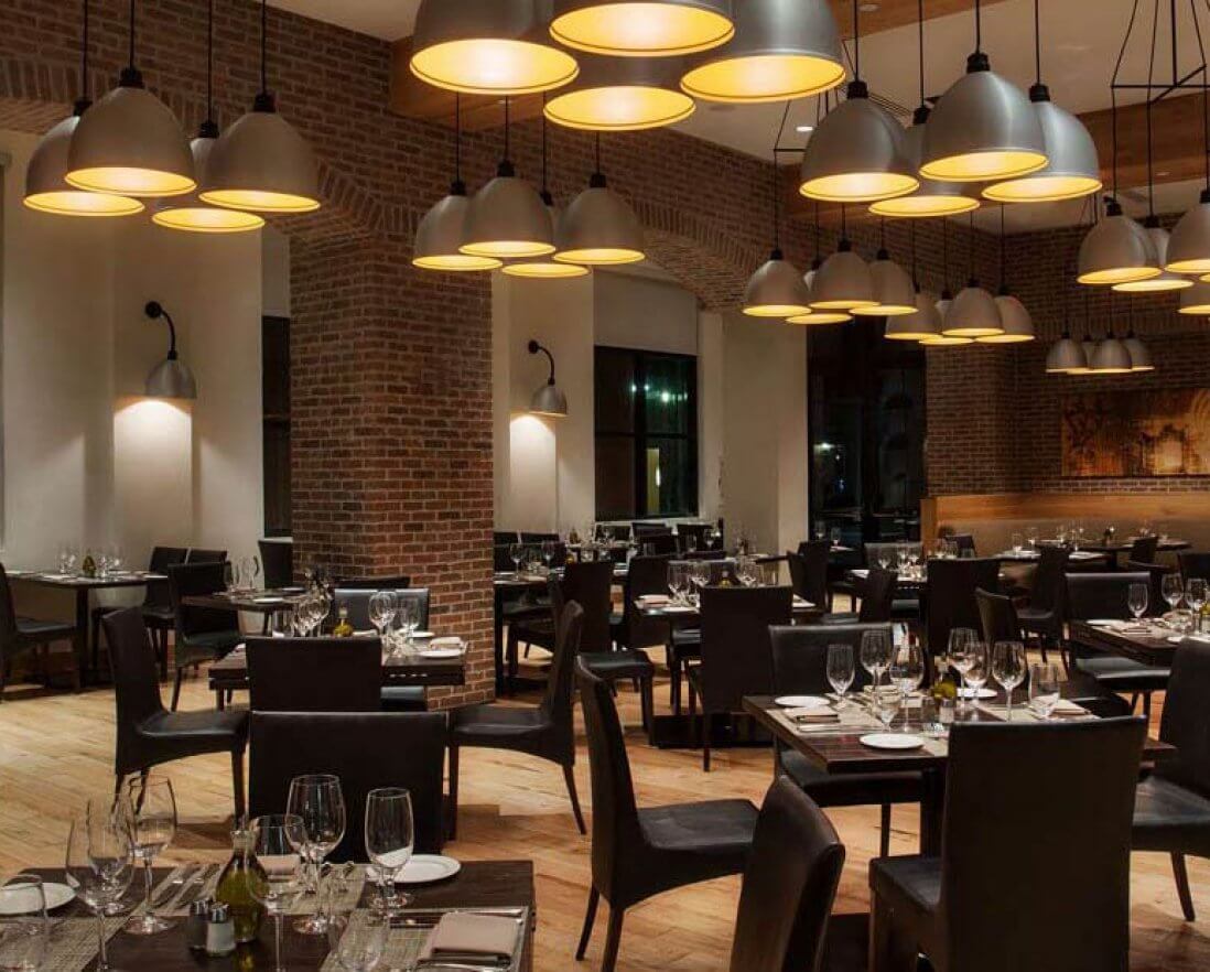 A modern, upscale restaurant with brick walls, wooden floors, dark tables and chairs, elegant table settings, and large pendant lights hanging from the ceiling. The space is empty and warmly lit.