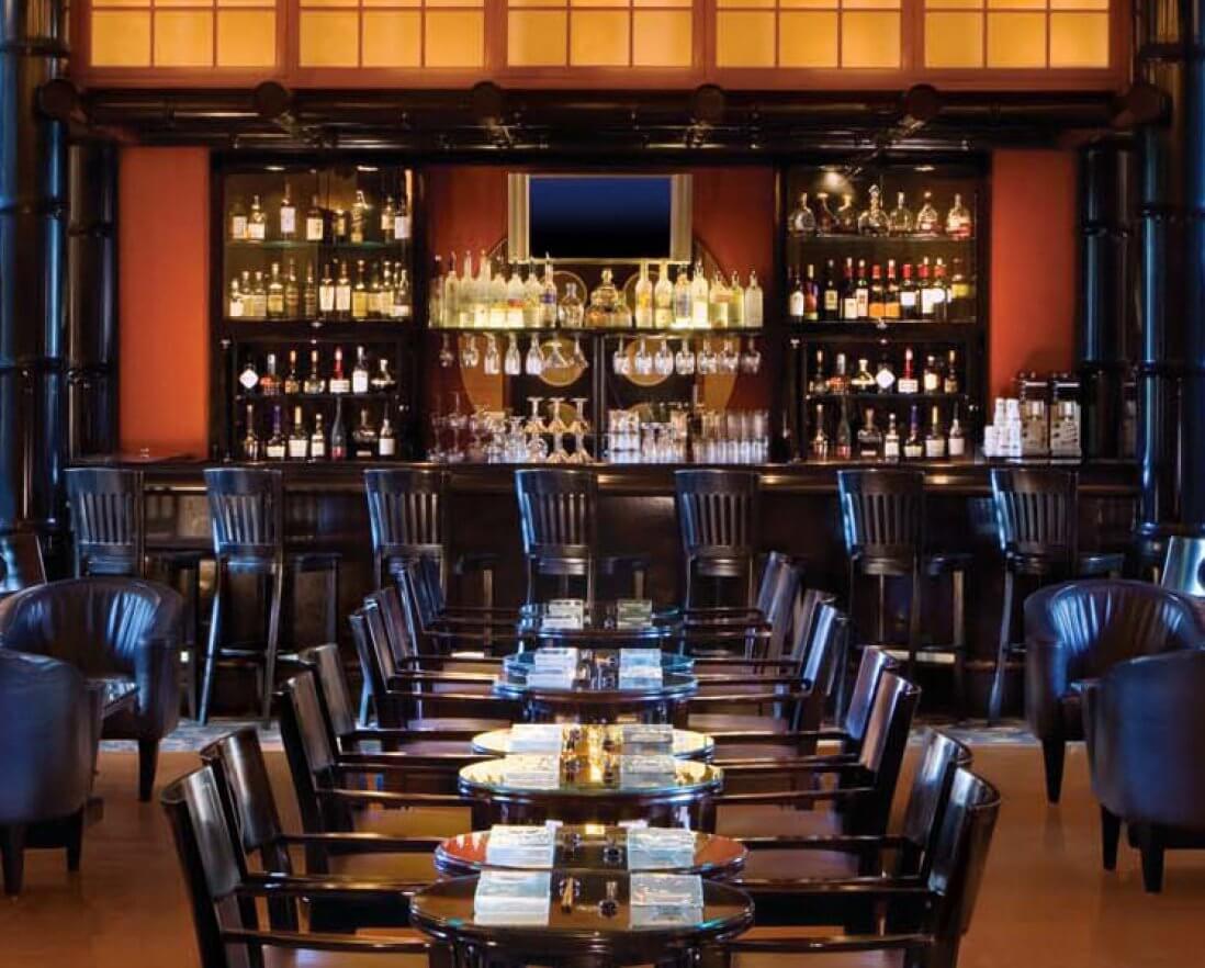 A stylish restaurant interior with black chairs and tables arranged facing a well-lit bar that features glass shelves with bottles and glassware. Warm wood and dark accents create an elegant, inviting atmosphere.