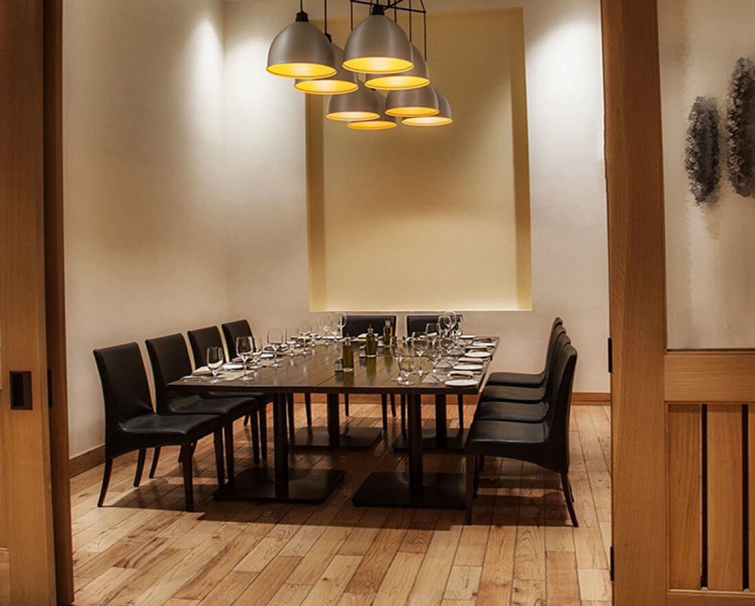 A modern private dining room with a long table set for twelve, surrounded by black chairs, wooden floors, soft lighting, and minimalist wall decor.