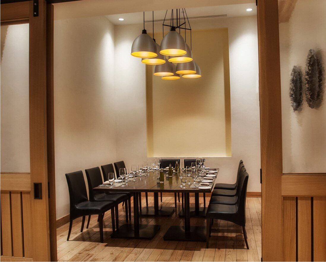 A modern, private dining room with a large rectangular table set for twelve, black chairs, wooden floors, and soft overhead pendant lights. The walls are minimal with subtle decor.