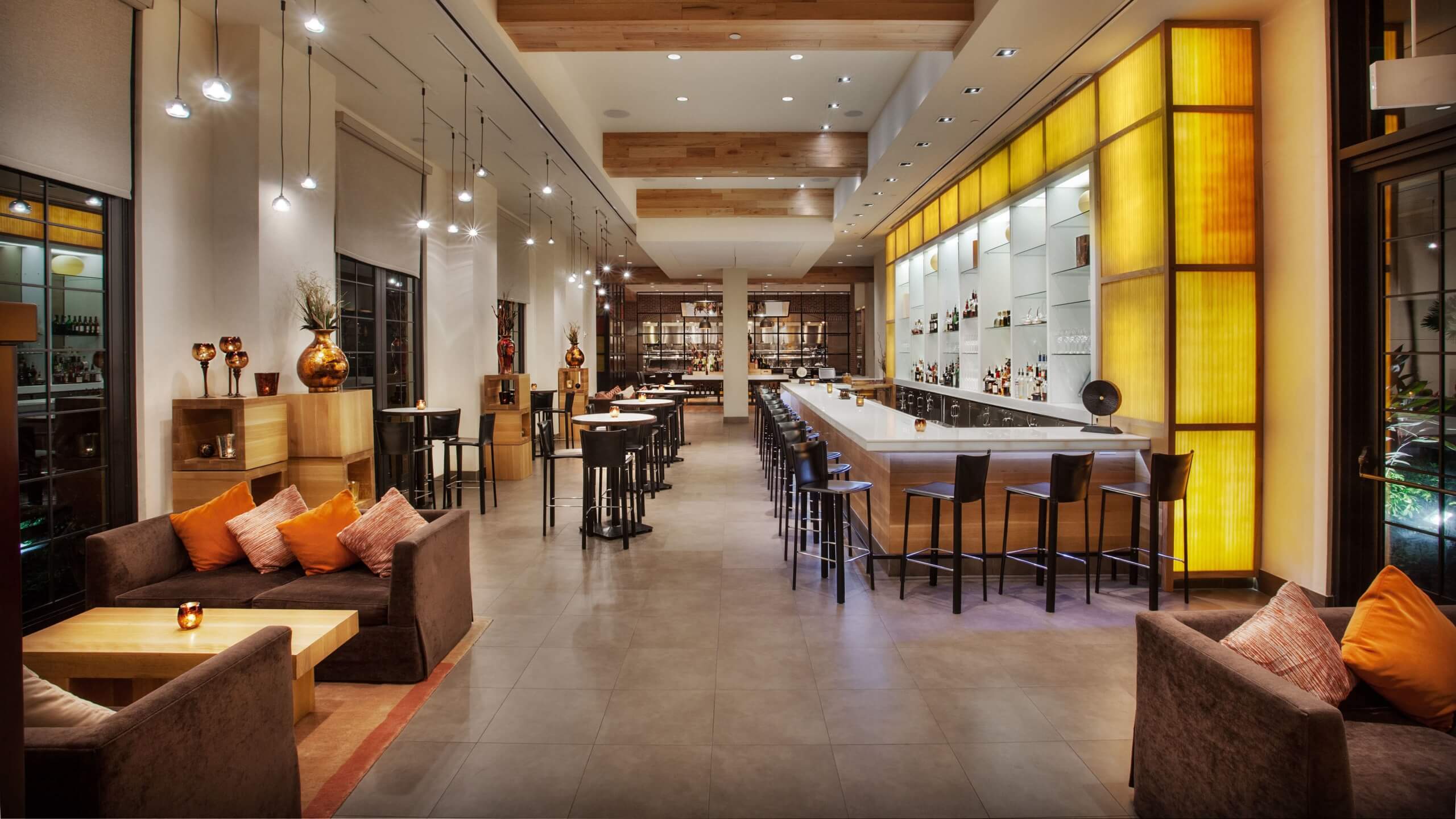 Modern restaurant interior with warm lighting, a long bar with high chairs, cozy seating areas with sofas and orange pillows, and decorative shelves lining the walls. Tables are neatly arranged throughout the spacious room.