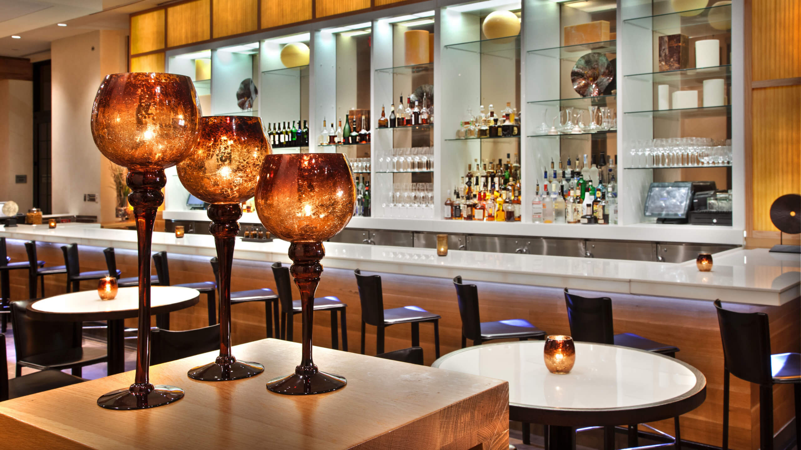 Modern bar interior with tall amber glass candle holders in the foreground, round white tables, black chairs, and a well-stocked backlit bar with shelves of bottles and glassware in the background.