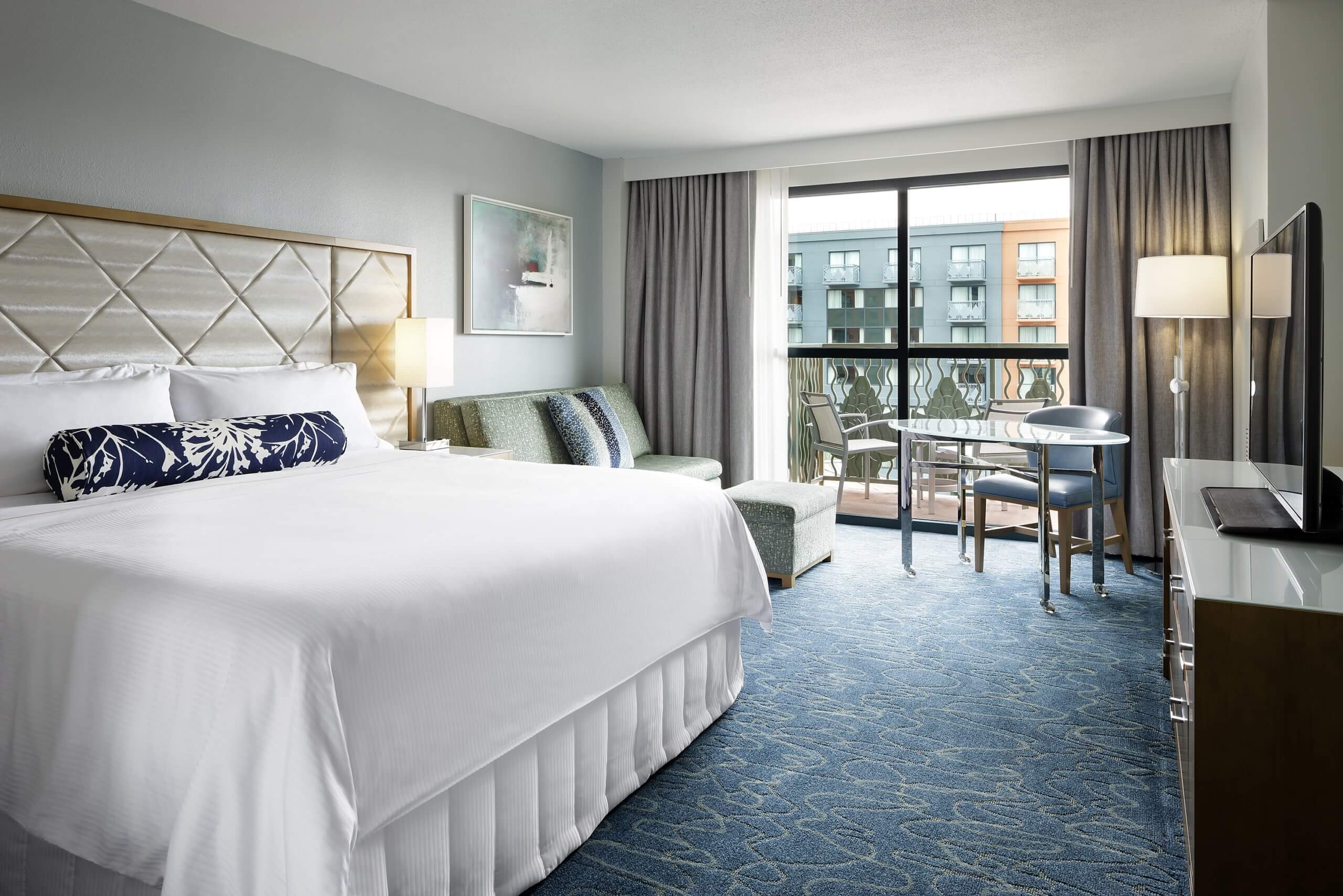 Bright hotel room with a large bed, blue carpet, a small seating area, balcony with chairs, and a view of another building through glass doors. Modern decor with soft colors and natural light.