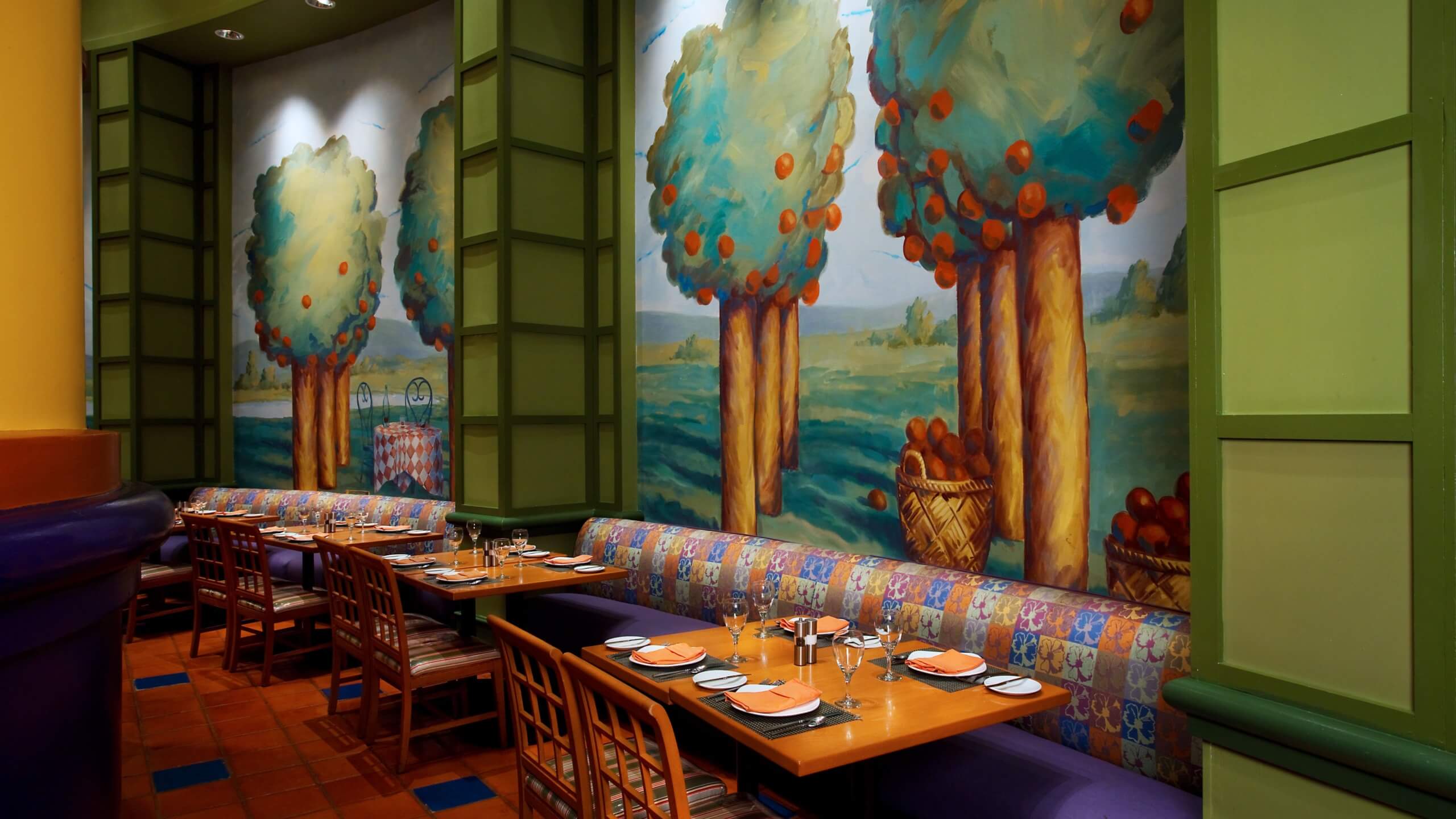 Une salle à manger de restaurant avec des tables et des chaises en bois, prête pour un repas. Le mur est orné d'une peinture murale colorée représentant des arbres avec des fruits orange et des paniers, créant ainsi une atmosphère fantaisiste inspirée de la nature.