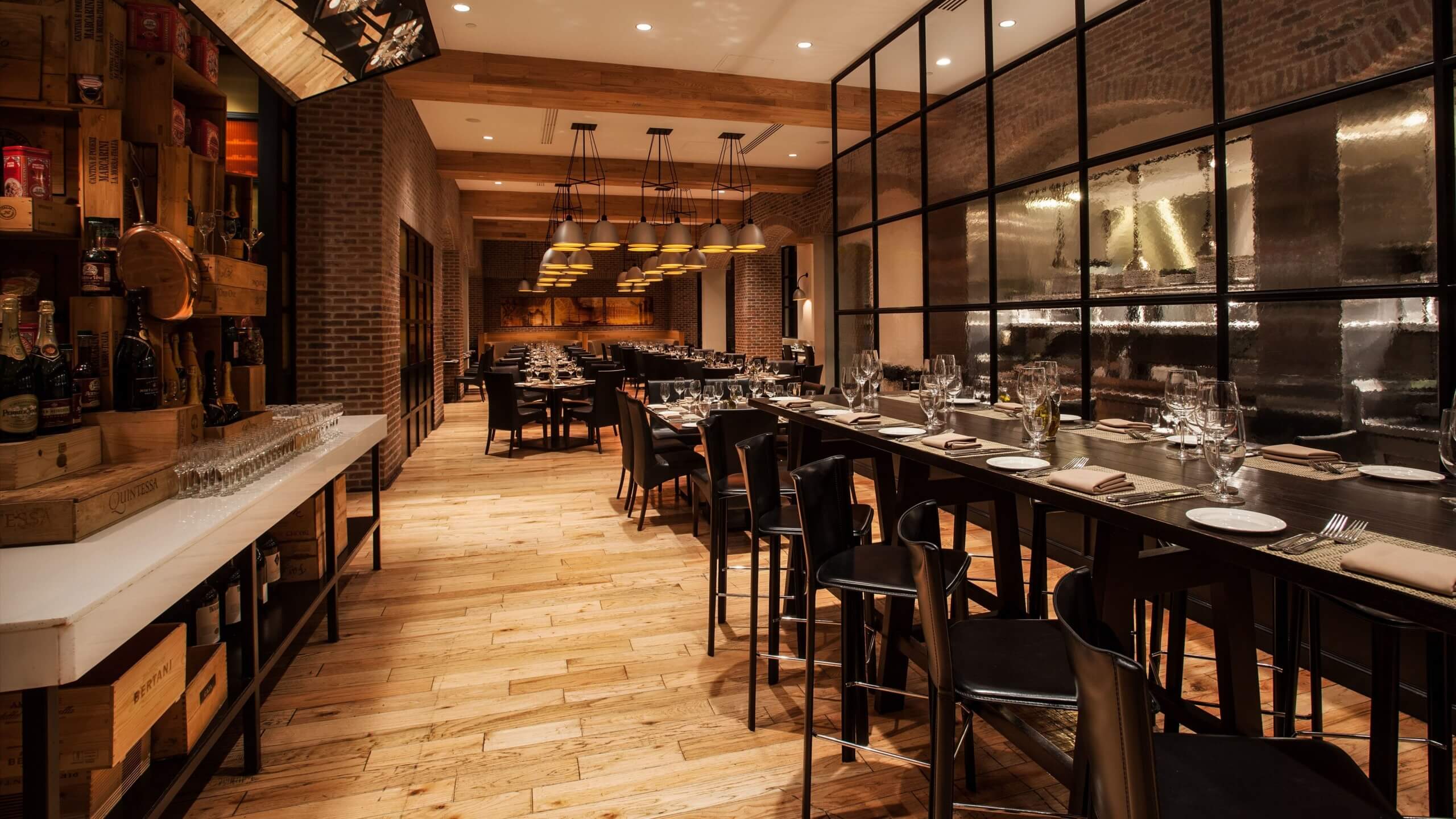 A stylish, warmly-lit restaurant with wooden floors, brick walls, and long black tables set with glassware and plates; pendant lights hang from the ceiling, creating an inviting and modern atmosphere.
