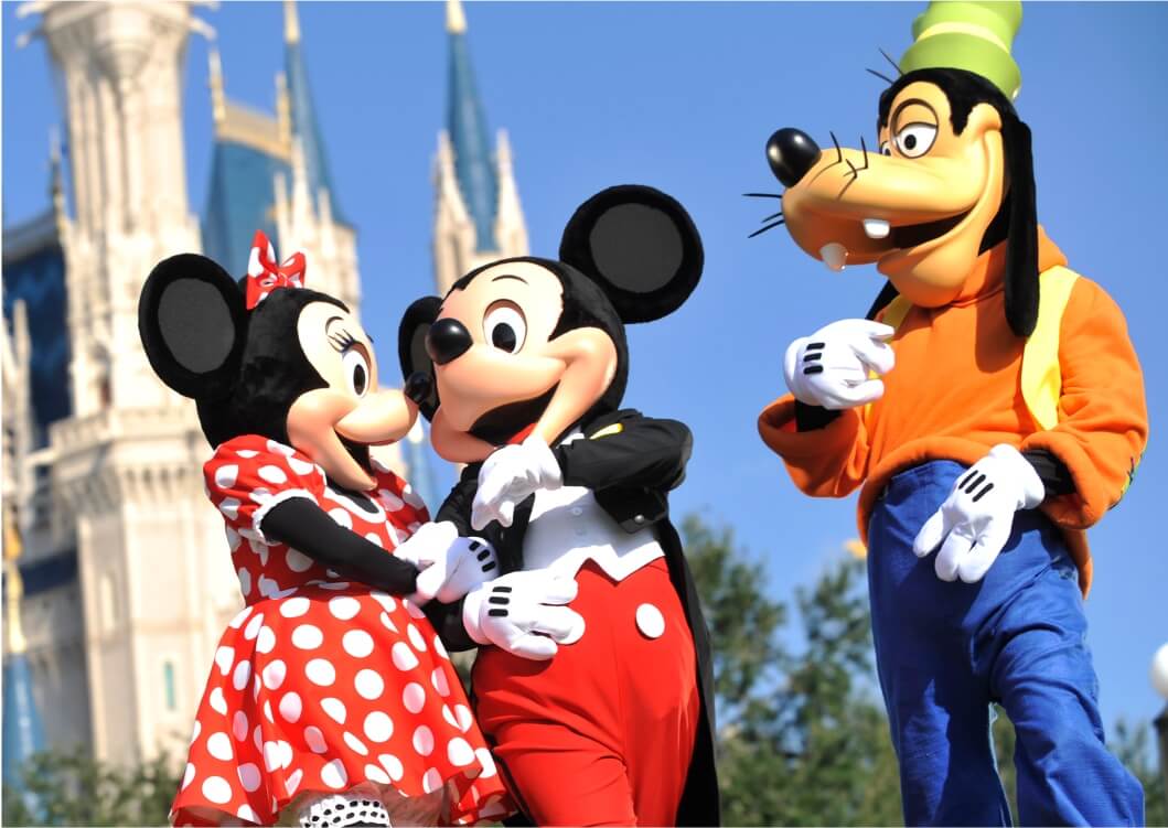 Minnie Mouse, Mickey Mouse, and Goofy, all in costume, stand together smiling in front of a castle at a Disney theme park, with clear blue sky in the background.