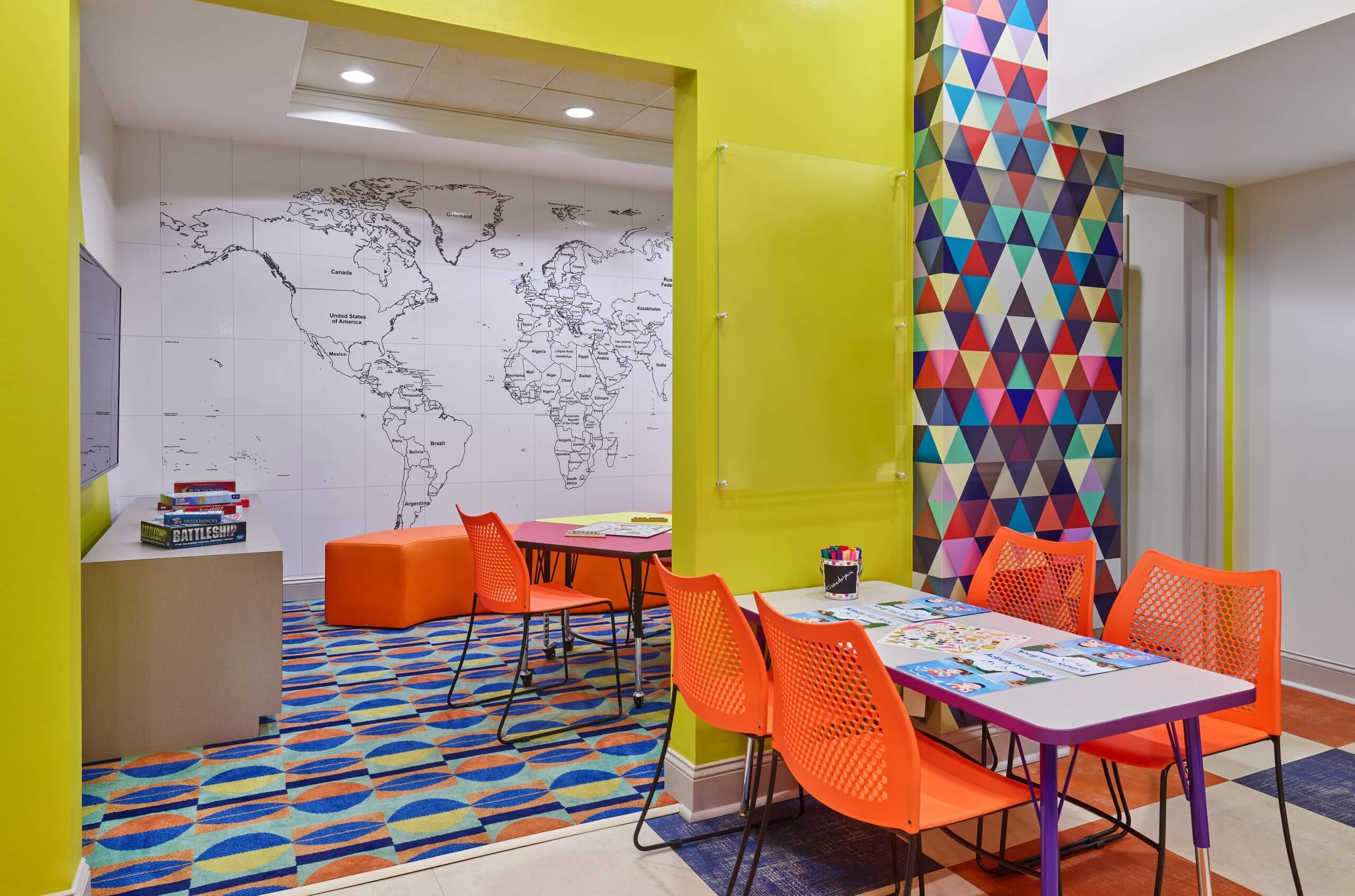 A colorful classroom with orange chairs, tables with art supplies, a wall map of the world, geometric patterned walls, board games, and bright decor.