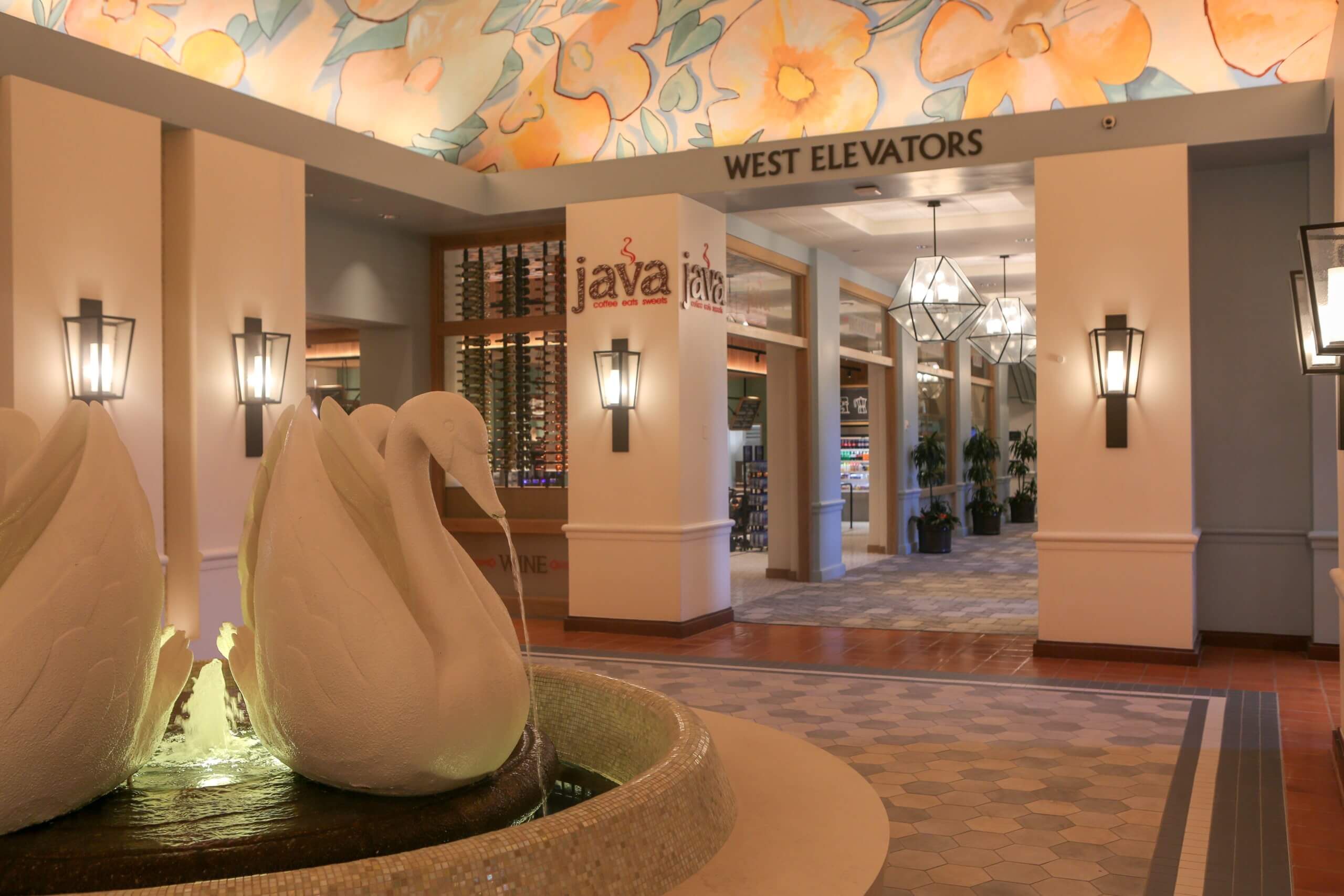 A hotel lobby features a white swan fountain in the foreground, floral ceiling art, warm lighting, a “Java” café sign, and a hallway leading to the west elevators.
