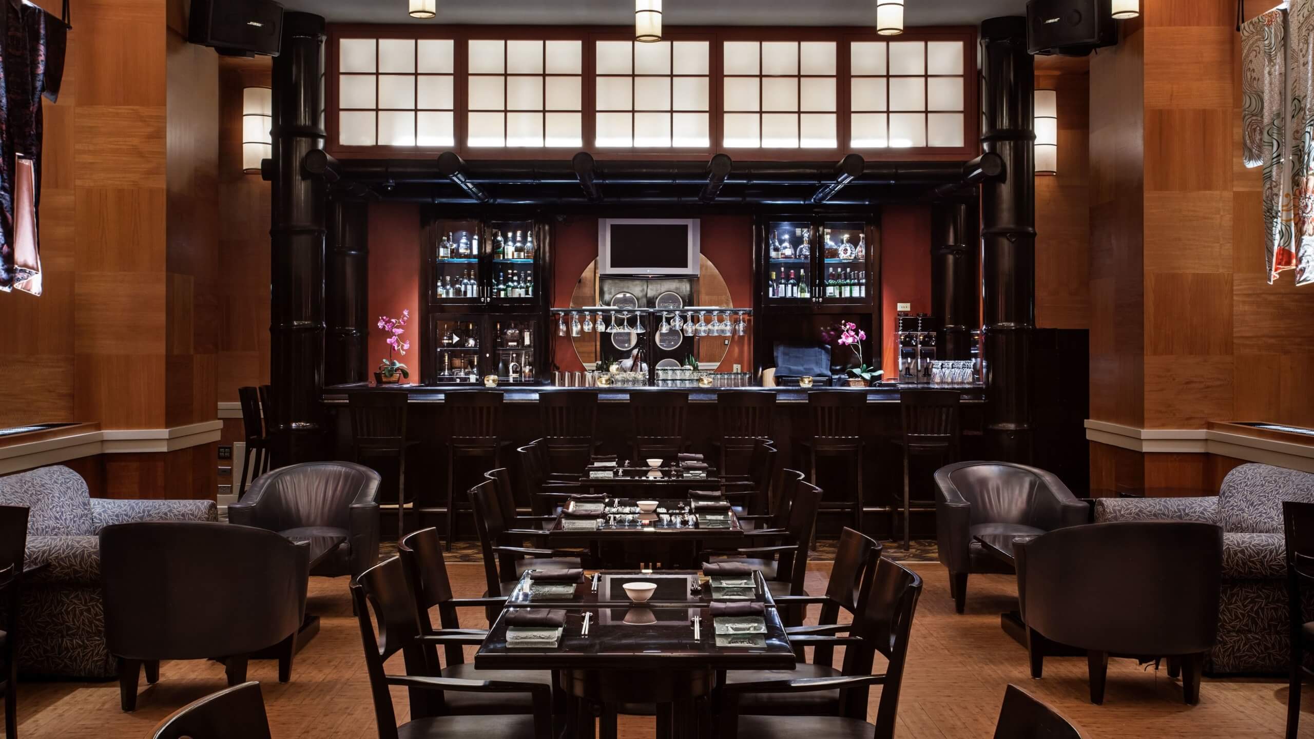 A modern restaurant interior with dark wood furniture, black chairs, neatly set tables, and a bar at the back displaying bottles and glasses. Soft lighting and decorative orchids create a warm, inviting atmosphere.