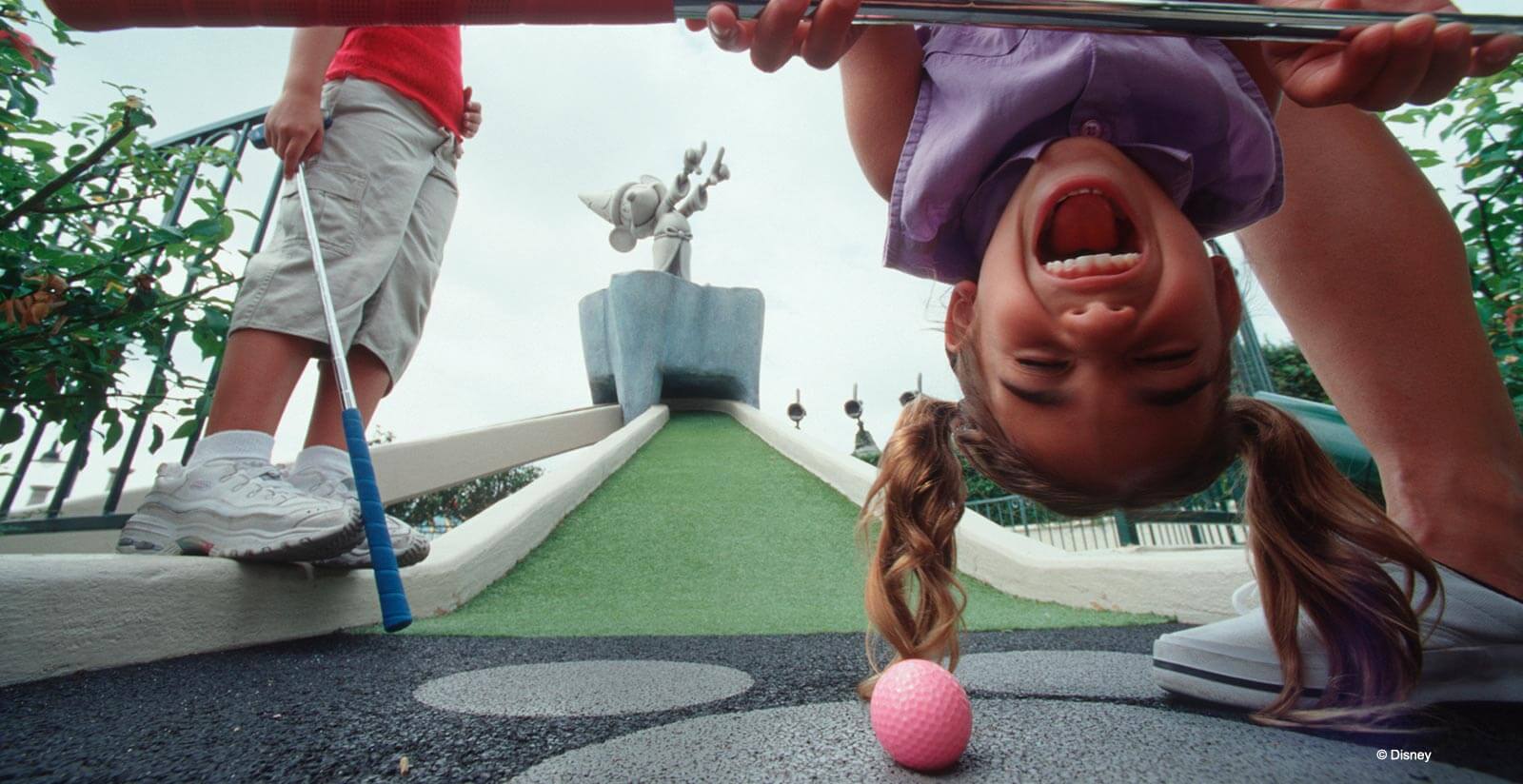 A young girl laughs while looking upside down at the camera on a mini-golf course, with a pink golf ball in front of her. Another person stands nearby holding a blue putter. A whimsical statue is in the background.