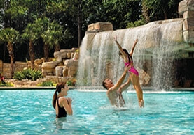 A man lifts a young girl in a pink swimsuit above the water in a swimming pool, while a woman watches nearby. A rocky waterfall and lush greenery are in the background.