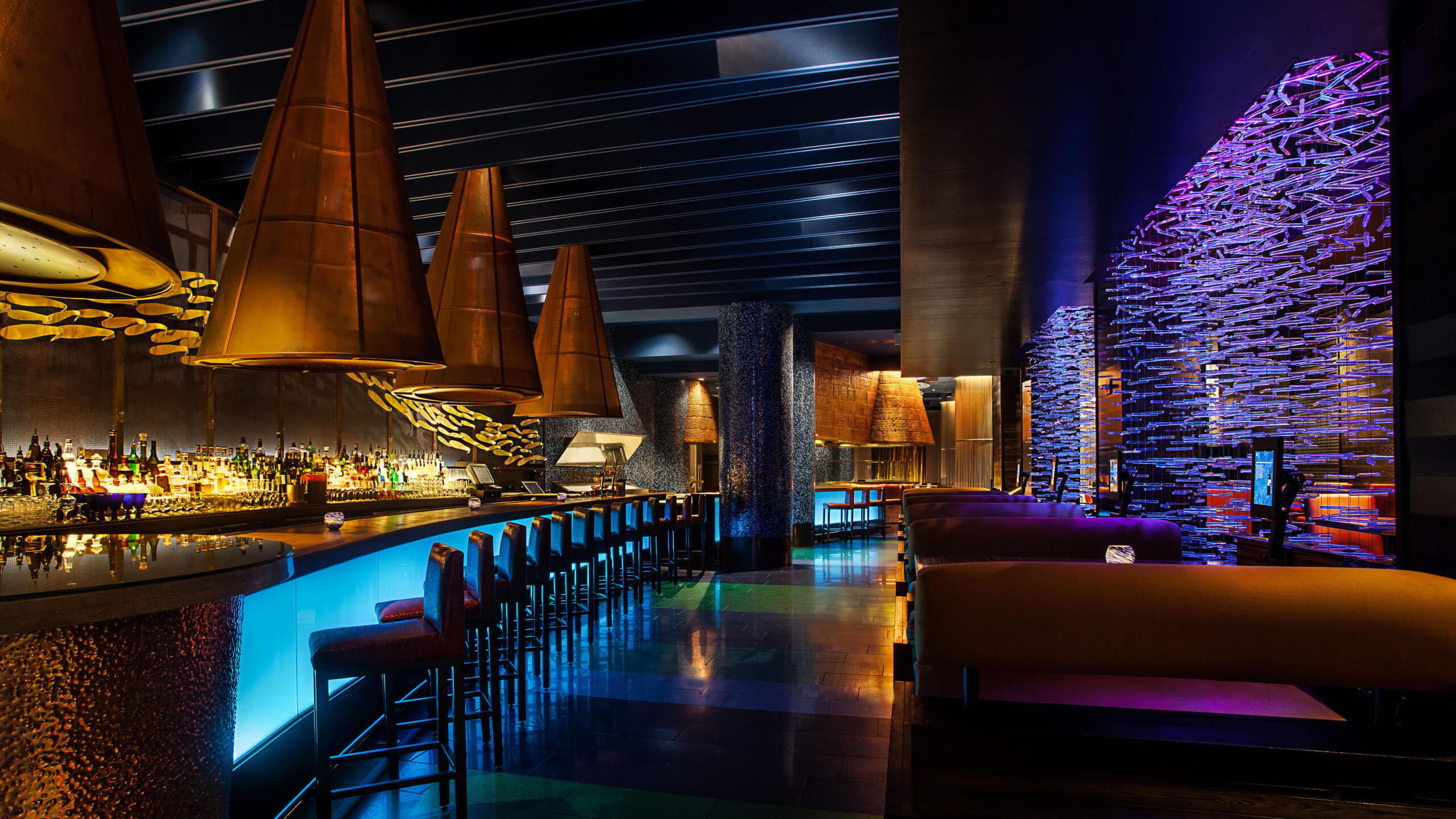 A modern, dimly lit bar with glowing blue counters, high-backed chairs, large copper pendant lights, and vibrant purple accent lighting along a textured wall, creating a stylish, upscale atmosphere.
