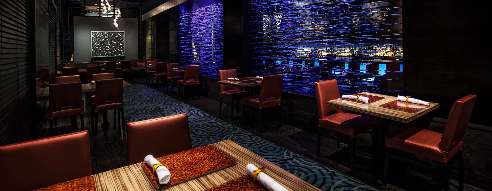 A modern restaurant with red chairs, patterned blue carpet, wooden tables set for dining, and a dramatic blue-lit textured wall creating a stylish ambiance.