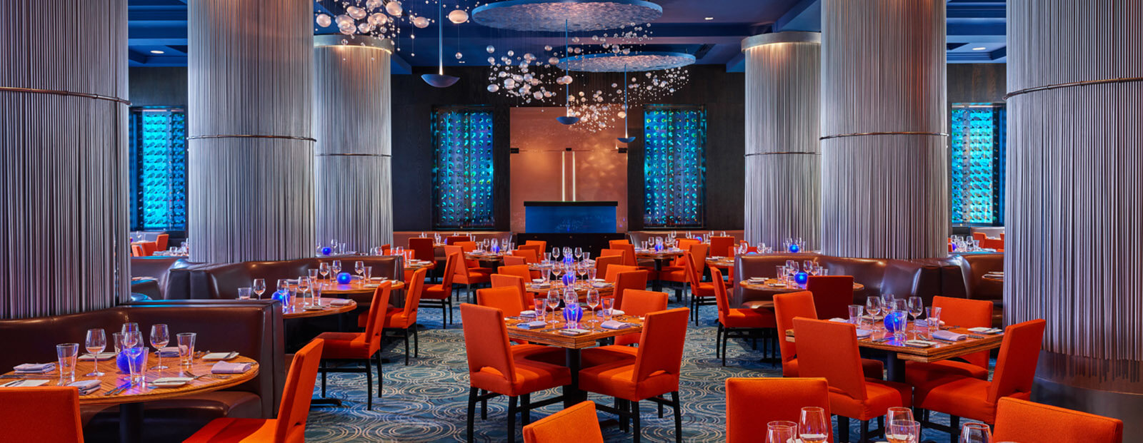 Elegant restaurant dining room with orange chairs, round wooden tables set with glassware and plates, soft blue lighting, modern chandeliers, and tall columns. Blue textured windows add a contemporary touch.