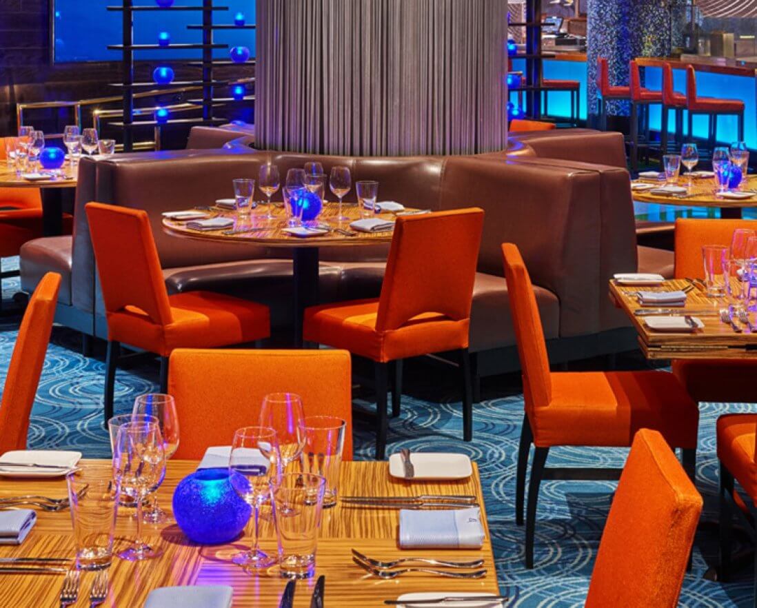 A modern restaurant dining area with orange chairs, wooden tables set with glasses and plates, blue glowing centerpiece lights, and stylish blue carpet flooring. Booth seating and ambient lighting create a vibrant, upscale atmosphere.
