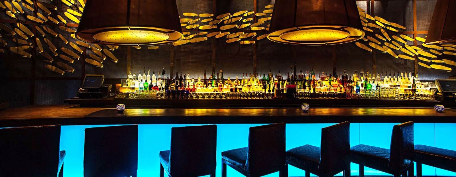 A modern bar with glowing blue counter lighting, dark barstools, and shelves stocked with bottles. Large pendant lights hang from the ceiling, and abstract golden wall art decorates the background.