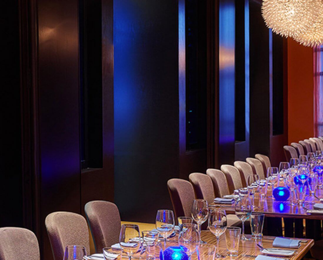 A modern dining room with a long wooden table set with glassware, napkins, and blue glowing centerpieces, surrounded by upholstered chairs and illuminated by spherical pendant lights.