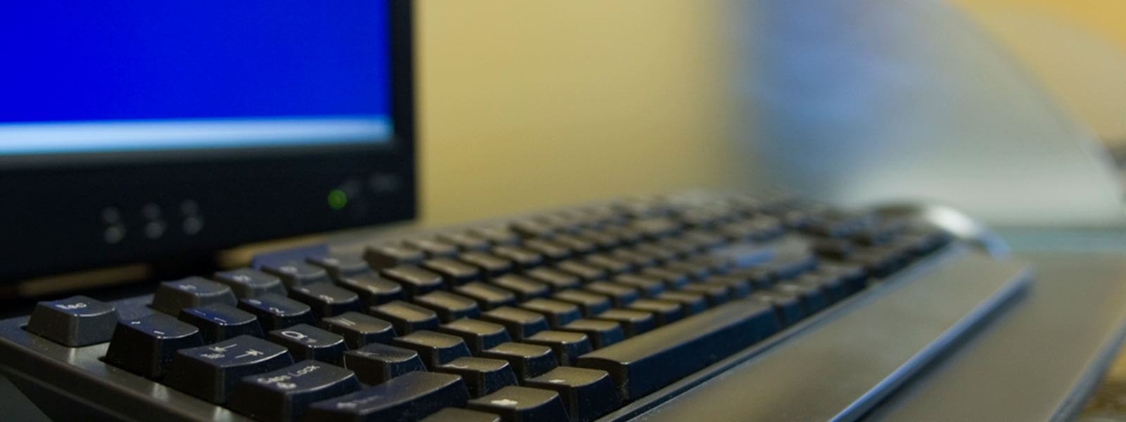A close-up view of a black computer keyboard in front of a monitor with a blue screen, both placed on a desk in an office or workspace setting.