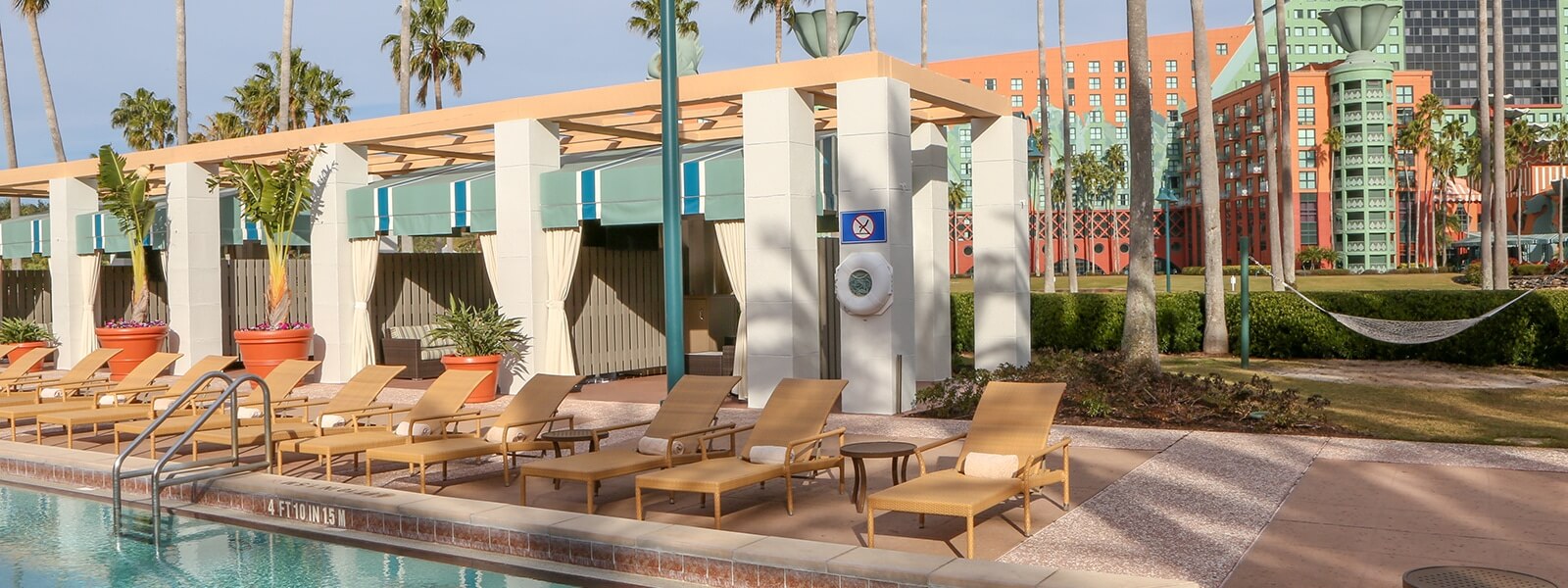 Lounge chairs line a poolside patio with large potted plants, cabanas, palm trees, and a building with colorful walls in the background; a hammock hangs among the trees.