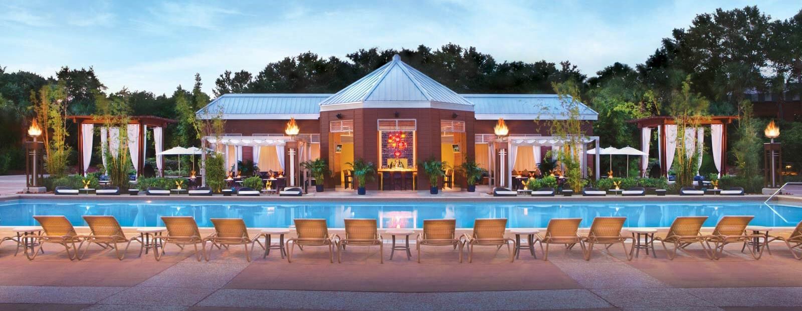 A large outdoor pool with rows of lounge chairs faces a modern clubhouse with a metal roof, surrounded by cabanas, tropical plants, and torches, set against a backdrop of trees at sunset.
