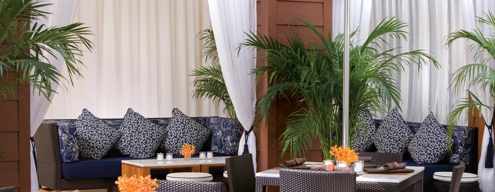Outdoor lounge area with a dark wicker sofa, blue and white patterned pillows, a table with orange flowers, chairs, green potted plants, and white curtains creating a relaxing, tropical atmosphere.