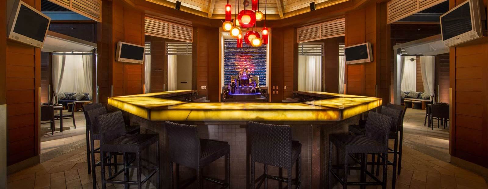 A modern, upscale bar with a glowing yellow countertop, dark barstools, hanging red pendant lights, and a vibrant, illuminated back shelf with bottles. Private seating areas are visible in the background.