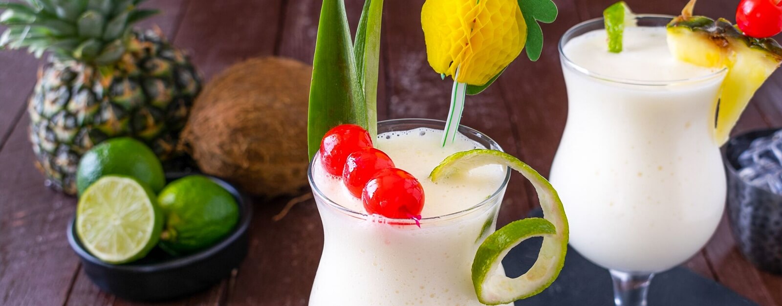 Two glasses of piña colada garnished with cherries, pineapple leaves, and decorative picks; fresh pineapple, coconut, and limes are in the background on a wooden table.