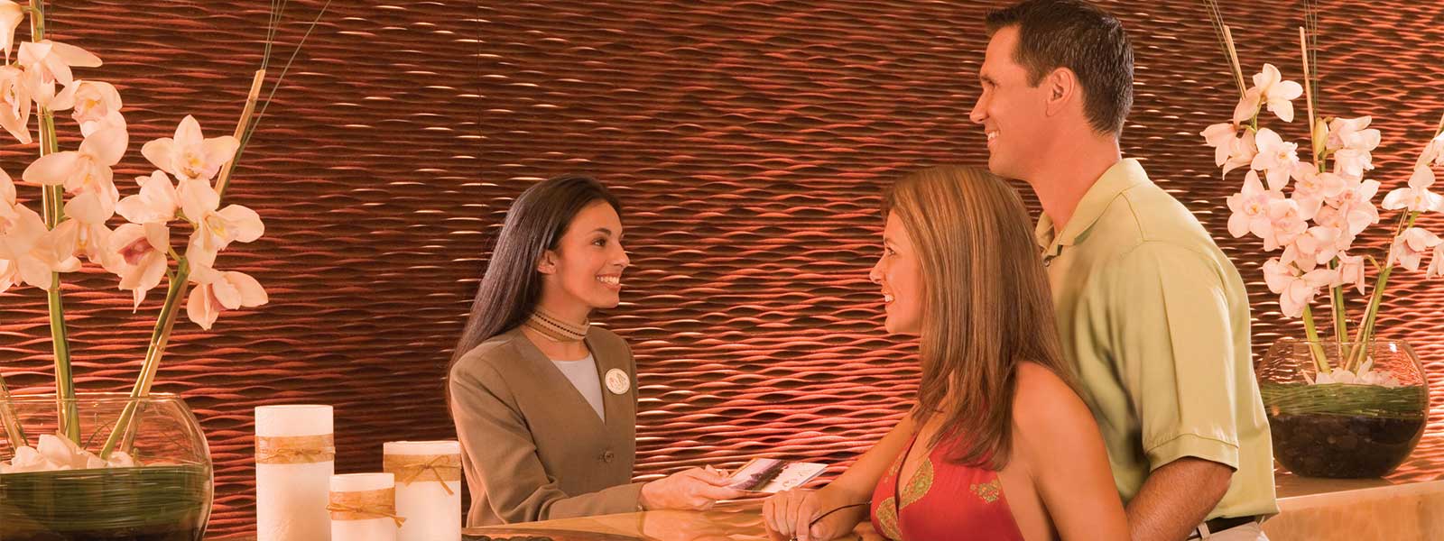 A smiling hotel receptionist hands key cards to a couple at the front desk, with decorative orchids and a textured wall in the background.
