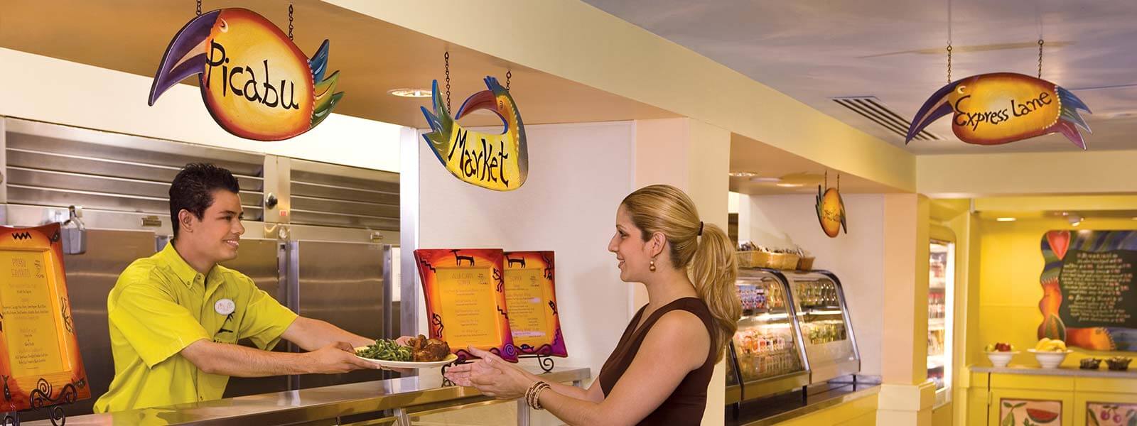 A smiling server in a lime-green shirt hands a plate of food to a woman at a colorful cafeteria counter, with bright, decorative signs hanging above the serving area.