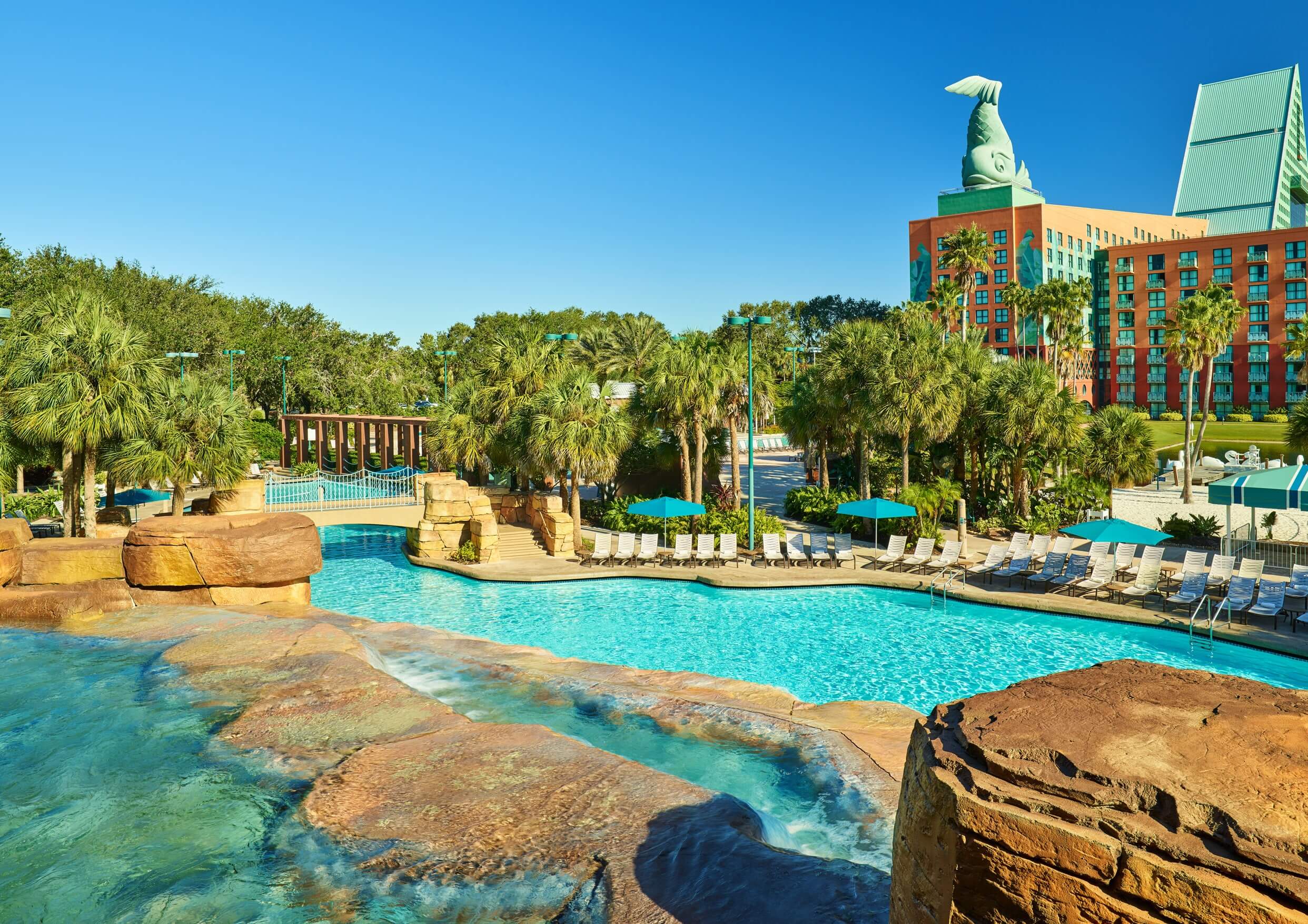 Large resort pool surrounded by palm trees and lounge chairs, with rock formations in the foreground and a colorful hotel with a decorative rooftop in the background under a clear blue sky.