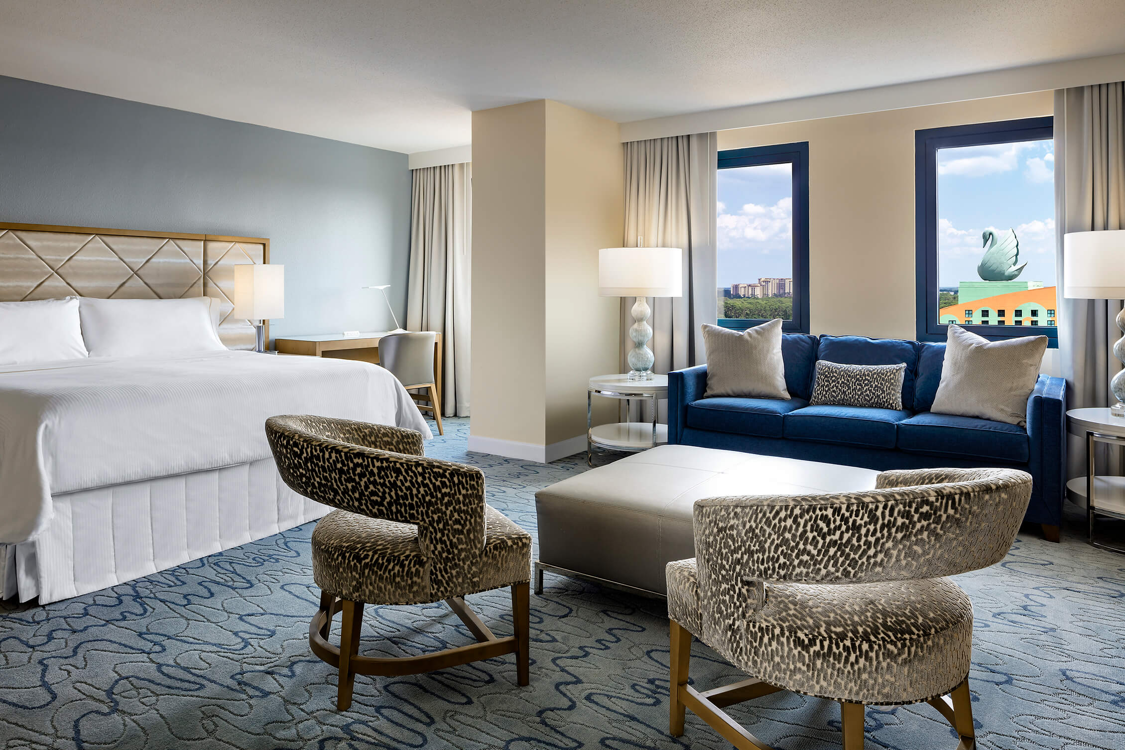 Spacious hotel room with king-size bed, two patterned armchairs, blue sofa, coffee table, desk, and large windows with a view of a cityscape and a dolphin statue outside.