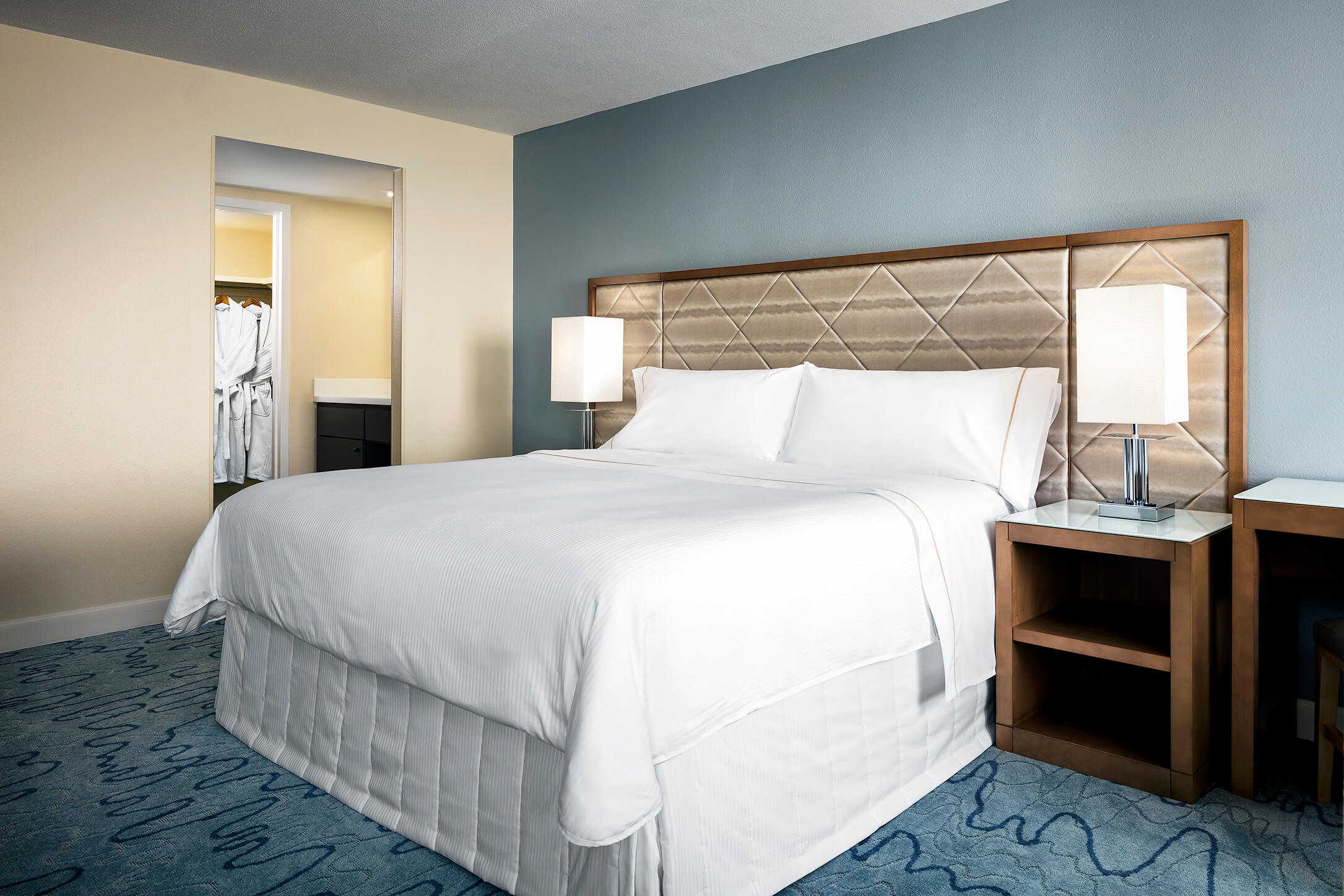 A neatly made hotel bed with white linens, flanked by two lamps on bedside tables, sits in a modern room. An open doorway reveals robes hanging in a closet area. The walls are light beige and blue.