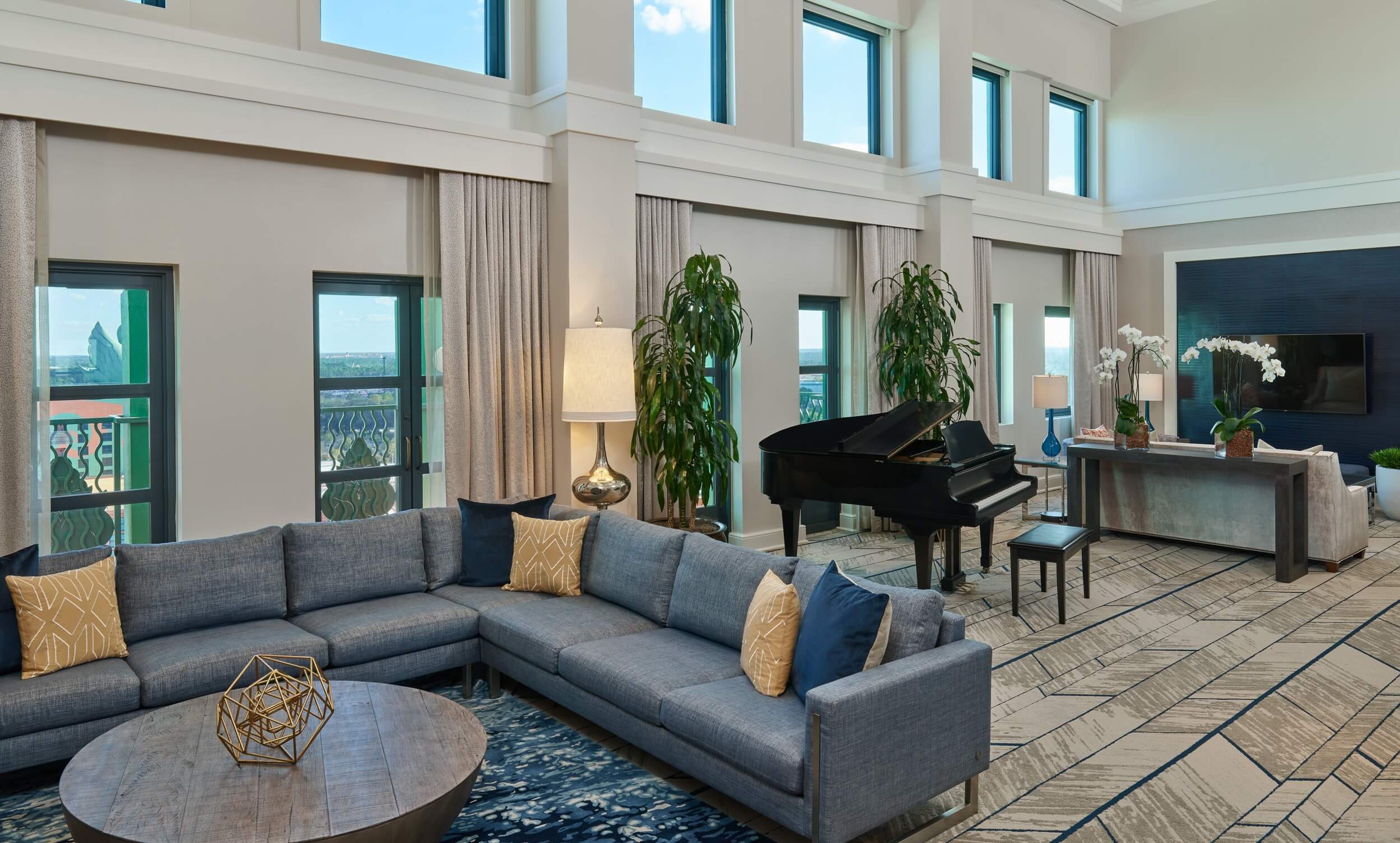 Spacious modern living room with tall windows, a large gray sectional sofa with yellow accent pillows, a round coffee table, a grand piano, indoor plants, and a TV console with decorative items.
