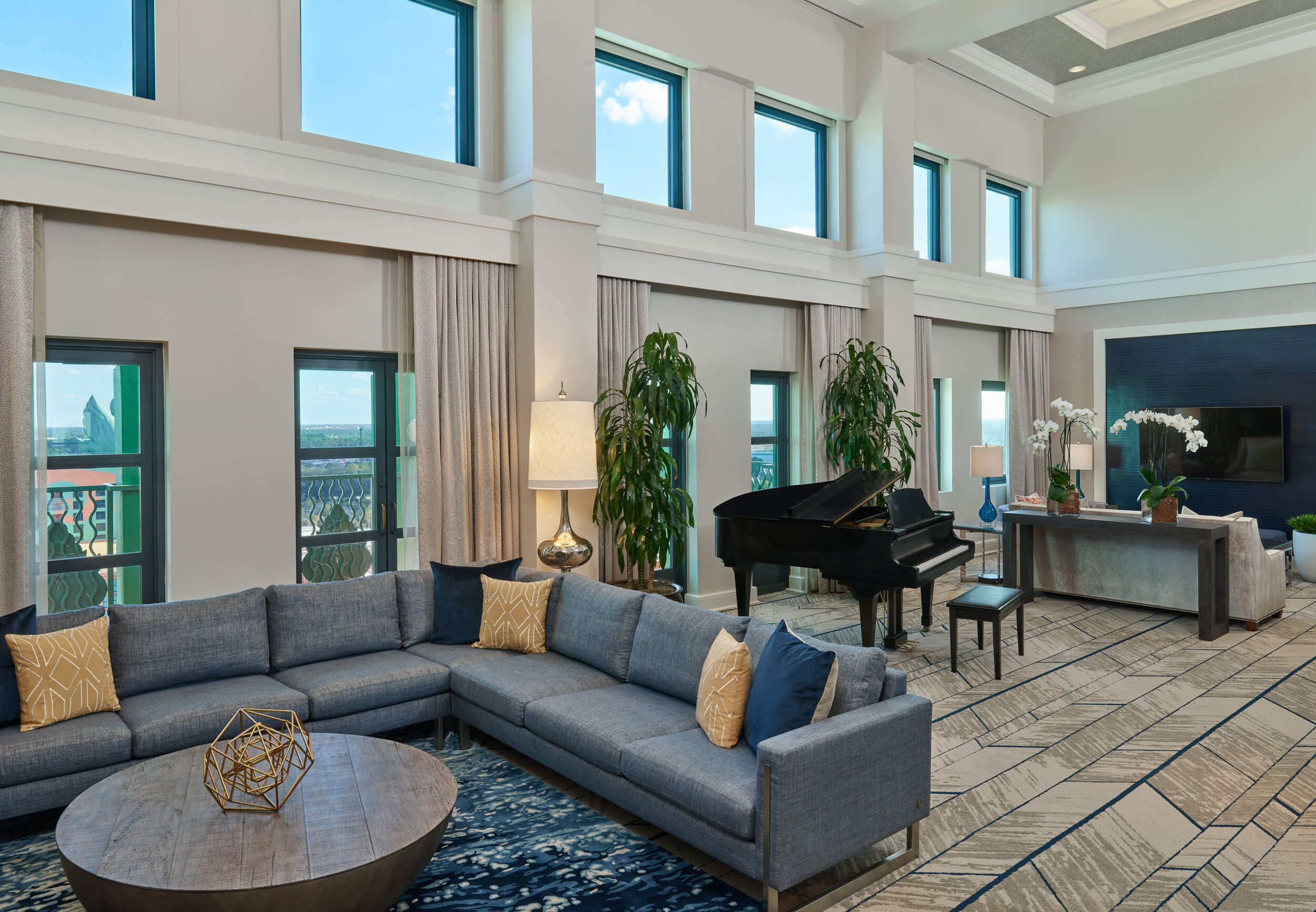 Spacious, modern living room with large windows, gray sectional sofa, round coffee table, black grand piano, tall plants, and TV on a console, decorated with yellow pillows and neutral tones.