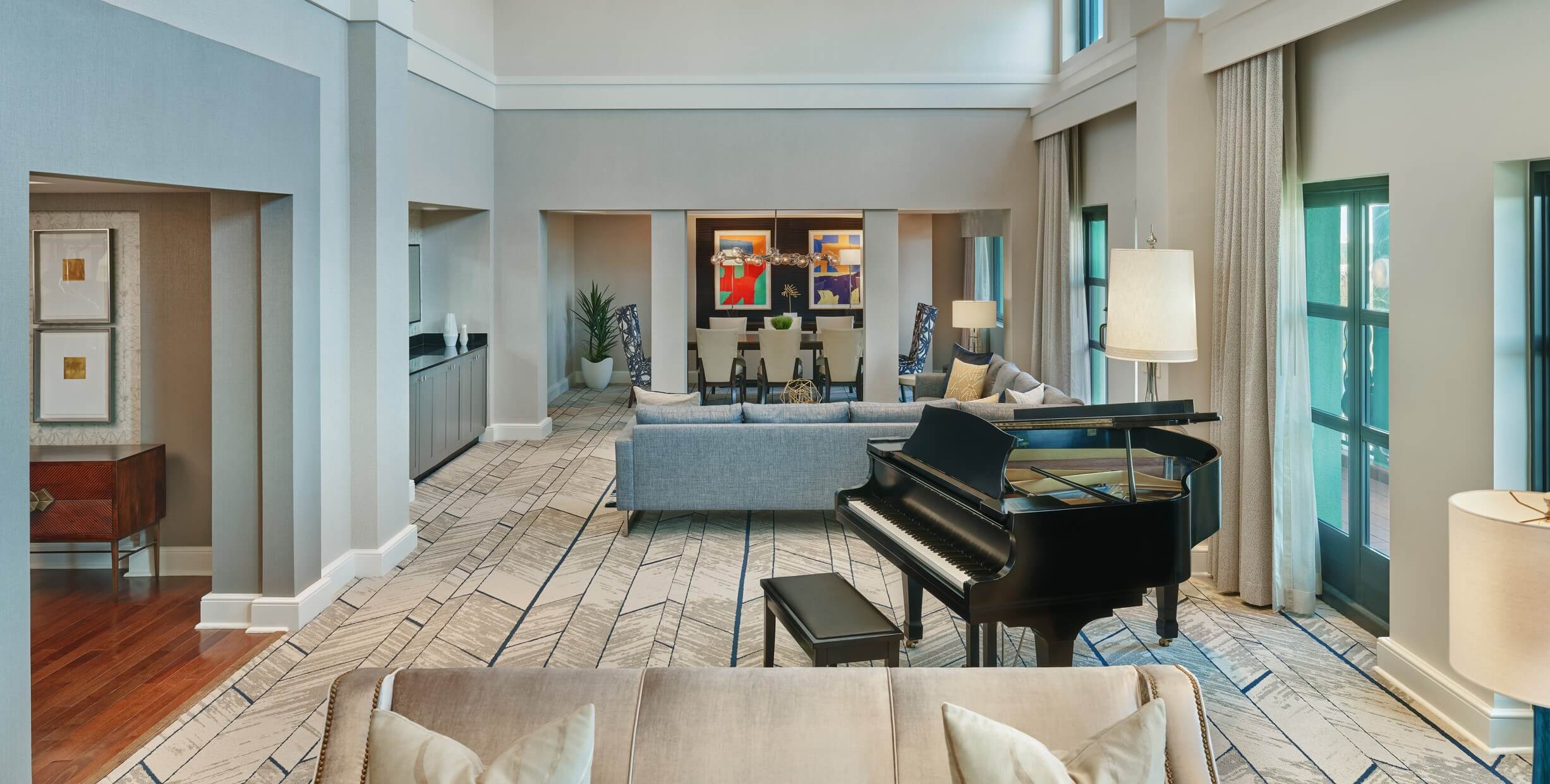 Spacious modern living room with high ceilings, large windows, a grand piano, neutral-toned sofas, patterned carpet, and contemporary decor. Artwork is displayed on the far wall. Natural light fills the space.
