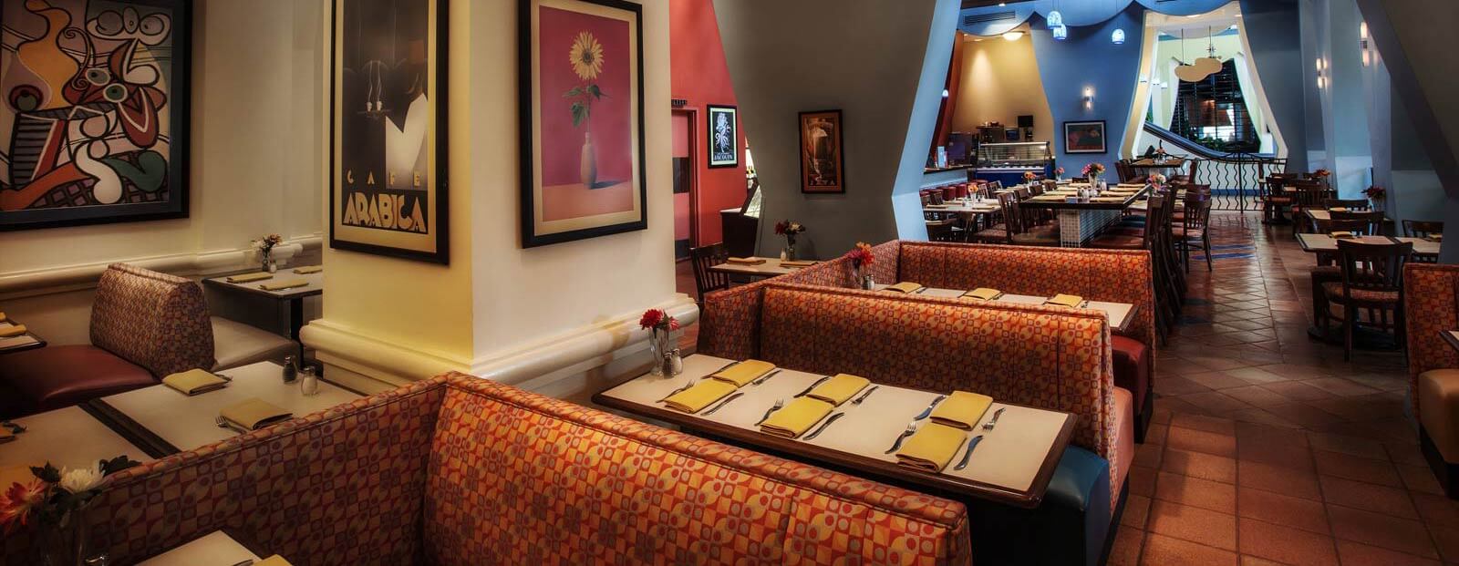 Colorful, modern restaurant interior with patterned booth seating, white tables set with yellow napkins, vibrant wall art, tiled floors, and a mix of booth and chair seating under warm lighting.