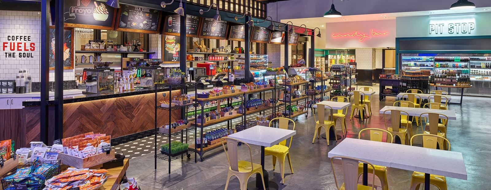 A modern café with colorful chairs and tables, a counter displaying snacks and drinks, overhead menu boards, and bright neon signs reading candy bar and pit stop in the background.