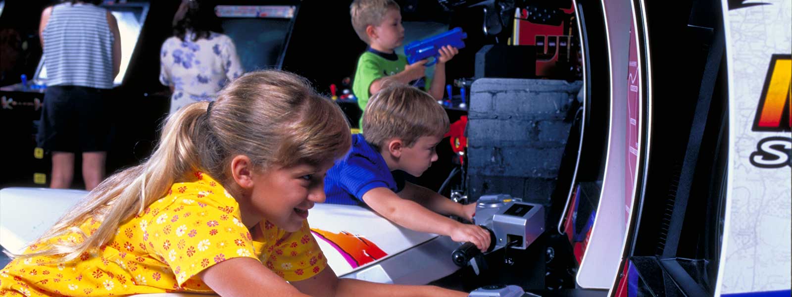 Children play arcade games, focusing on shooting-themed machines. A girl in a yellow shirt is in the foreground, while two boys play alongside her. The environment is colorful and lively.