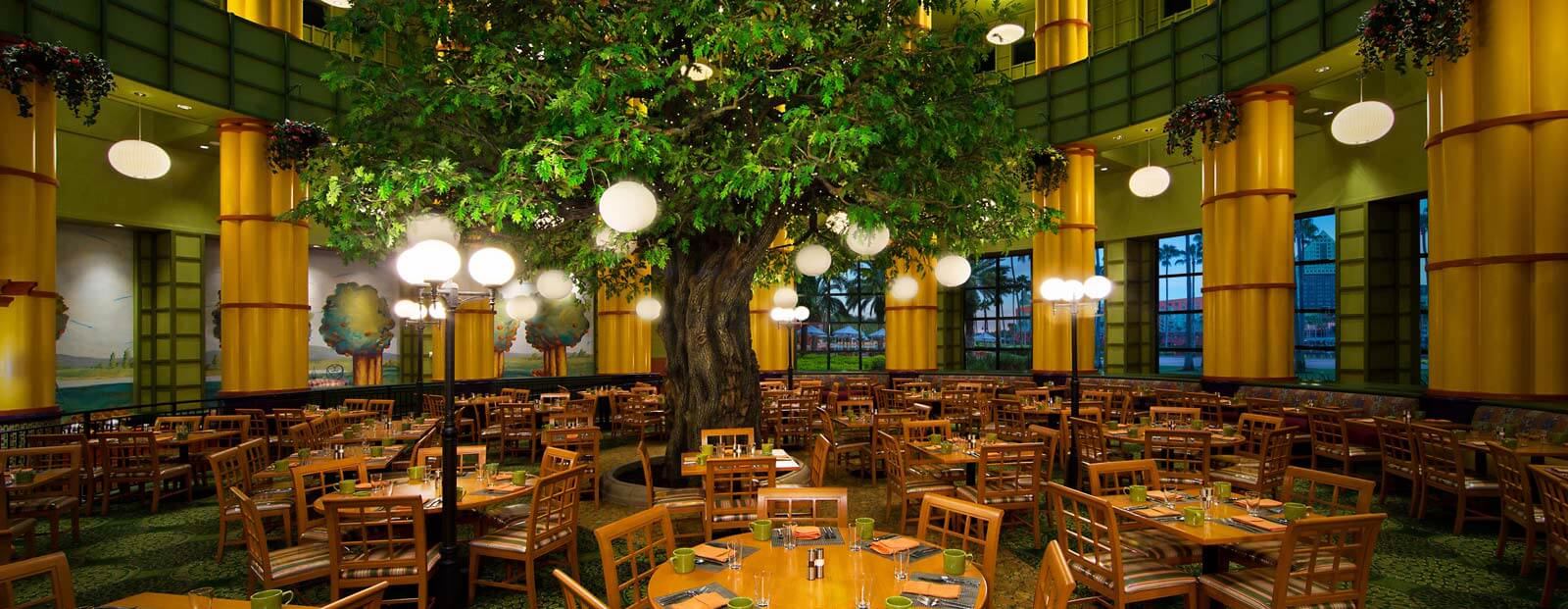 A spacious restaurant with wooden tables and chairs arranged around a large artificial tree in the center, glowing round light fixtures hanging from its branches, and tall yellow columns surrounding the room.