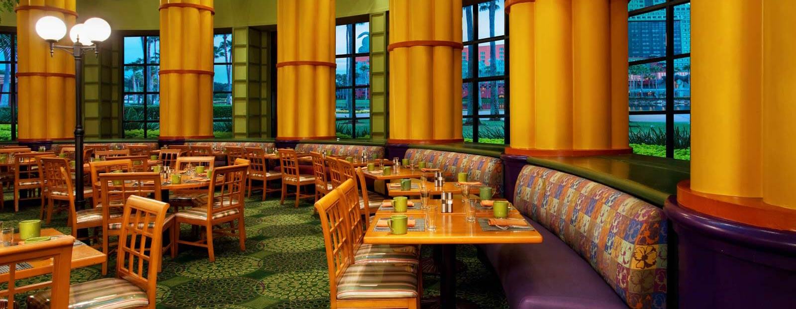 A brightly lit restaurant dining area with wooden tables and chairs, patterned booth seating, large yellow columns, and tall windows showing trees and buildings outside. Tables are set with plates, glasses, and condiments.