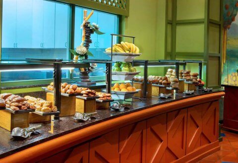 A breakfast buffet with various pastries, rolls, bananas, and fruit displayed on trays and stands on a long counter in a brightly lit room with green walls and a mural of trees.
