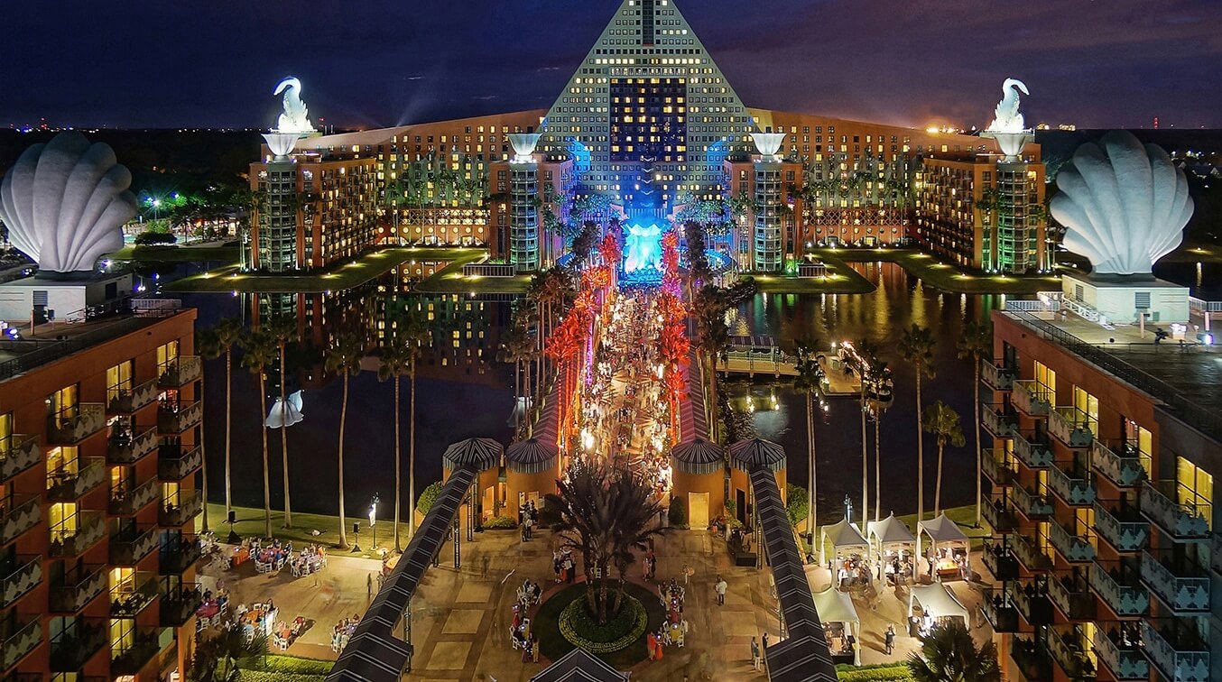 A nighttime view of a large, pyramid-shaped hotel with illuminated windows, flanked by two buildings with giant swan statues on top and palm trees lining a bustling central walkway filled with people and lights.