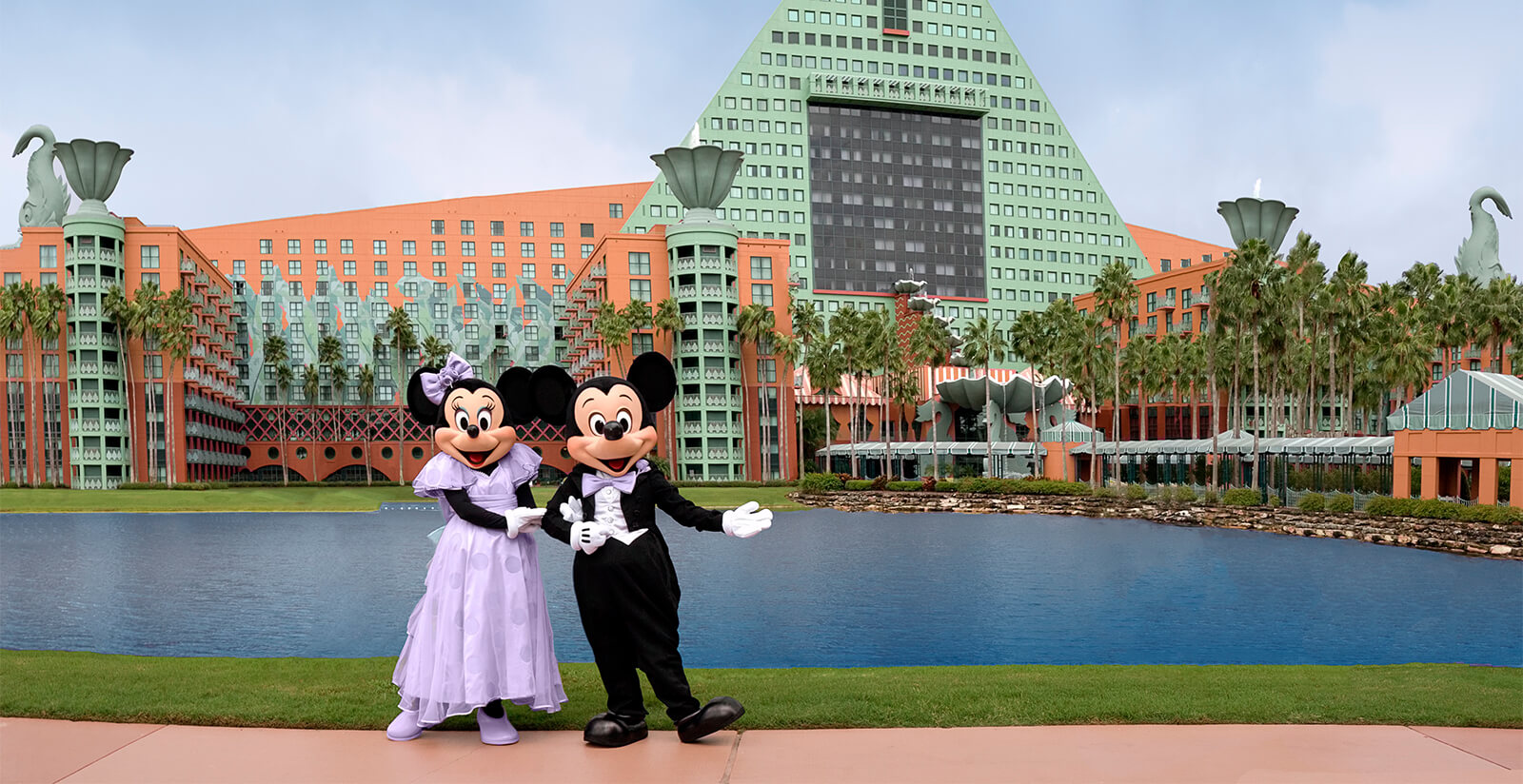 Minnie Mouse in a lavender dress and Mickey Mouse in a tuxedo pose together in front of a large lakeside hotel with green geometric roofs and palm trees in the background.