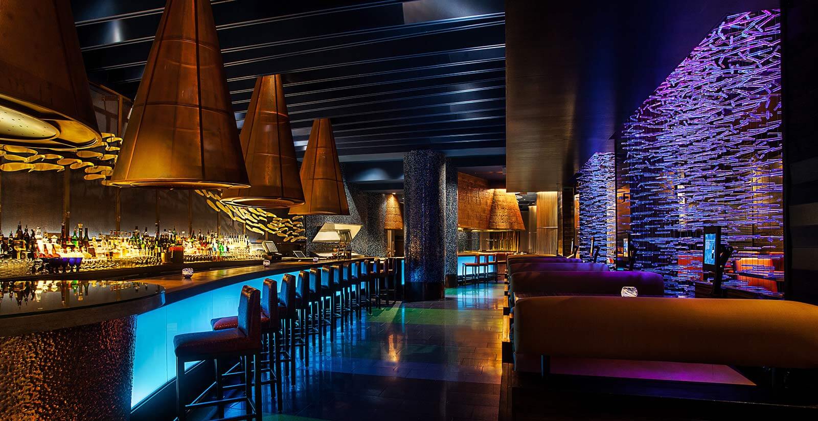 A modern, dimly lit bar with glowing blue counters, high stools, large cone-shaped copper light fixtures, and purple decorative wall accents, creating a sleek and vibrant atmosphere.
